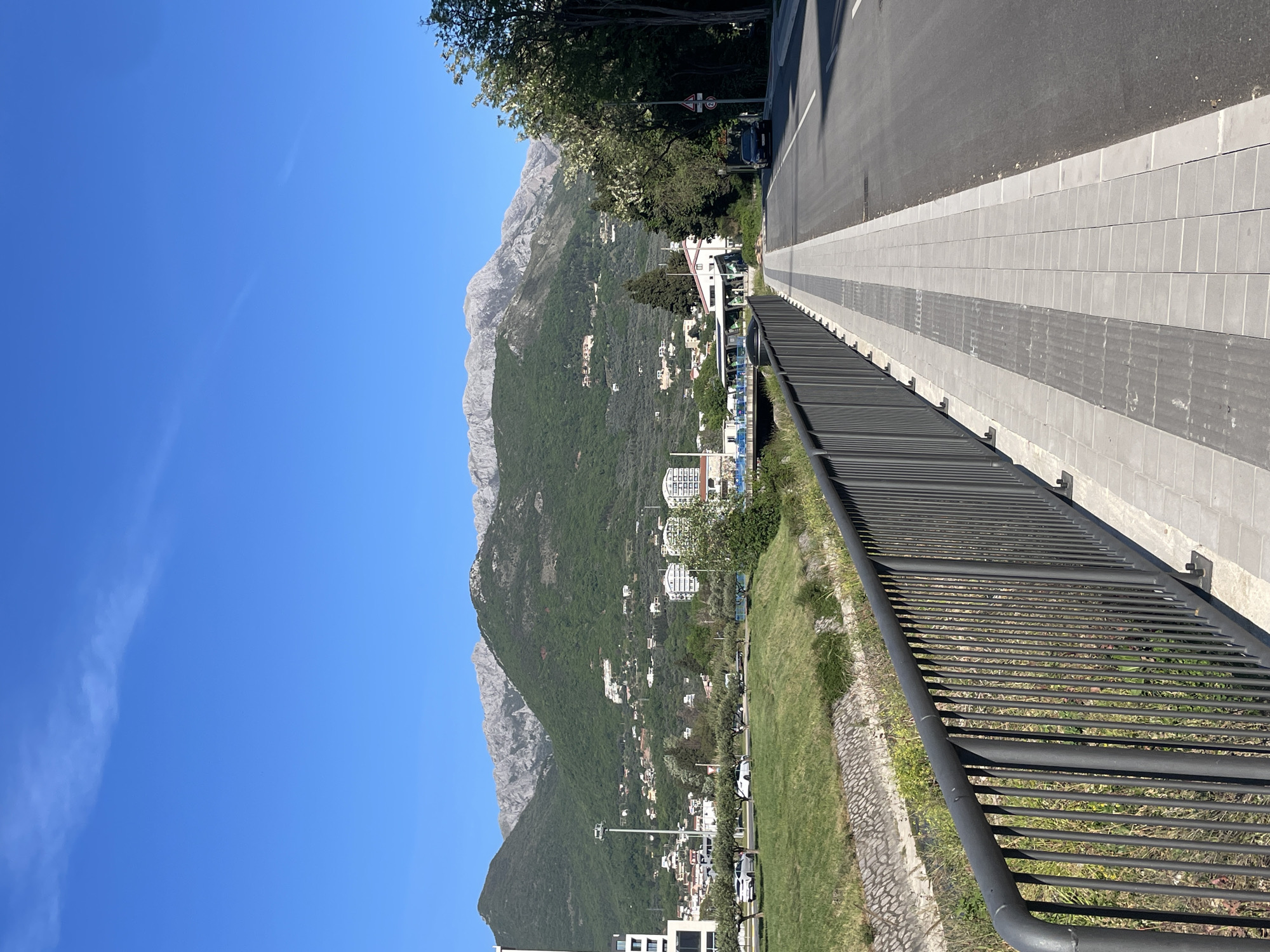 The image shows a view of the Dinaric Alps from the city of Bar, Montenegro. The foreground features a black metal railing with a grassy slope beneath it, separating it from the street. A sidewalk paved with gray rectangular tiles runs alongside the street. The street has a dark asphalt surface. Beyond the street, the landscape transitions to a hillside covered in green vegetation and scattered buildings. In the background, a mountain range rises, with rocky peaks and forested slopes, standing against a clear blue sky. No people are visible.