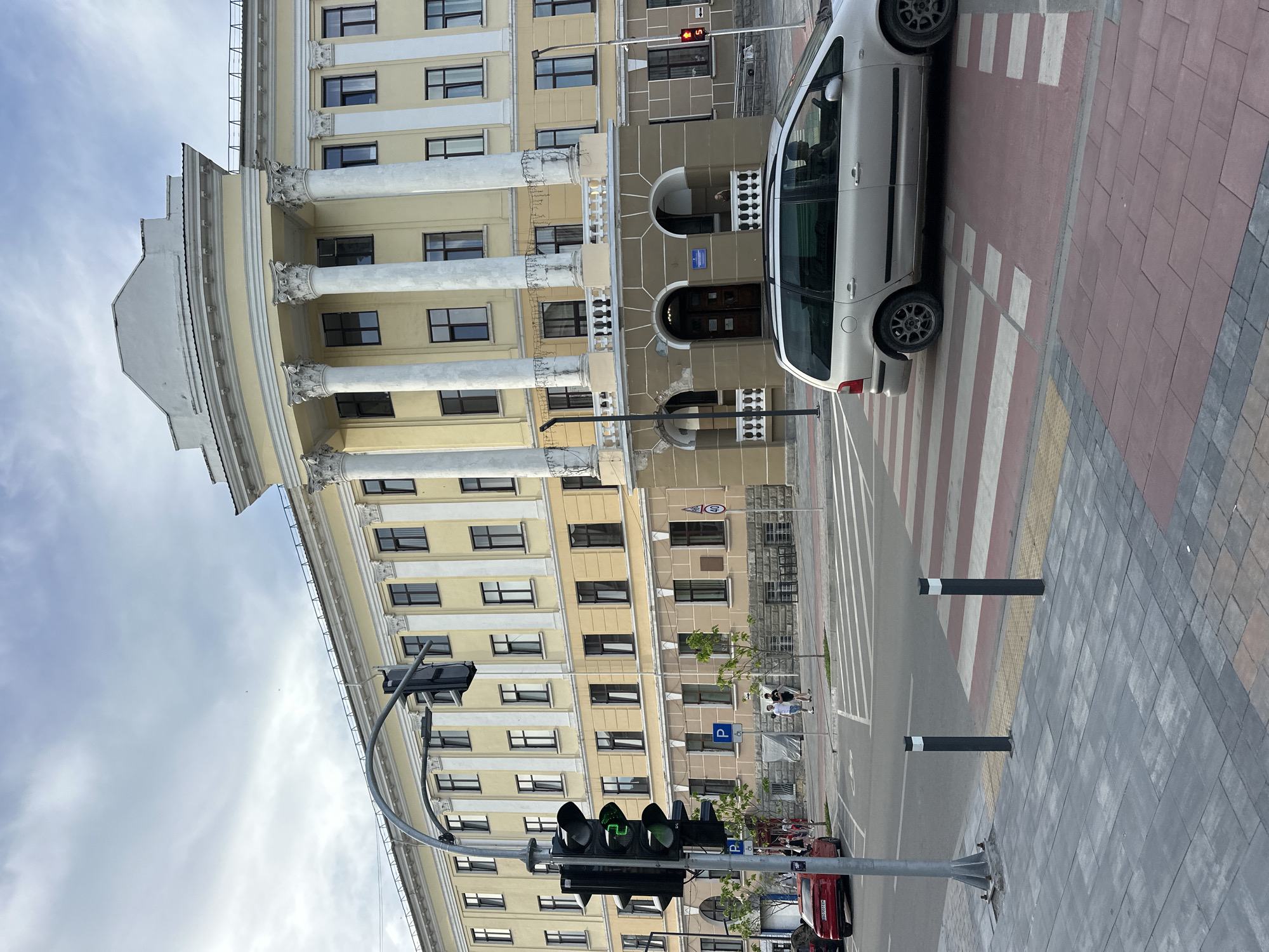 The image shows a street scene in Ternopil, Ukraine, featuring a prominent yellow building with classical architectural elements, including columns and a curved facade. In the foreground, a crosswalk with red and white stripes is visible, with a silver minivan crossing it. A traffic light is also present, displaying a green signal. On the sidewalk, several people are walking. The sky is overcast. The building appears to be an institutional or administrative structure.
