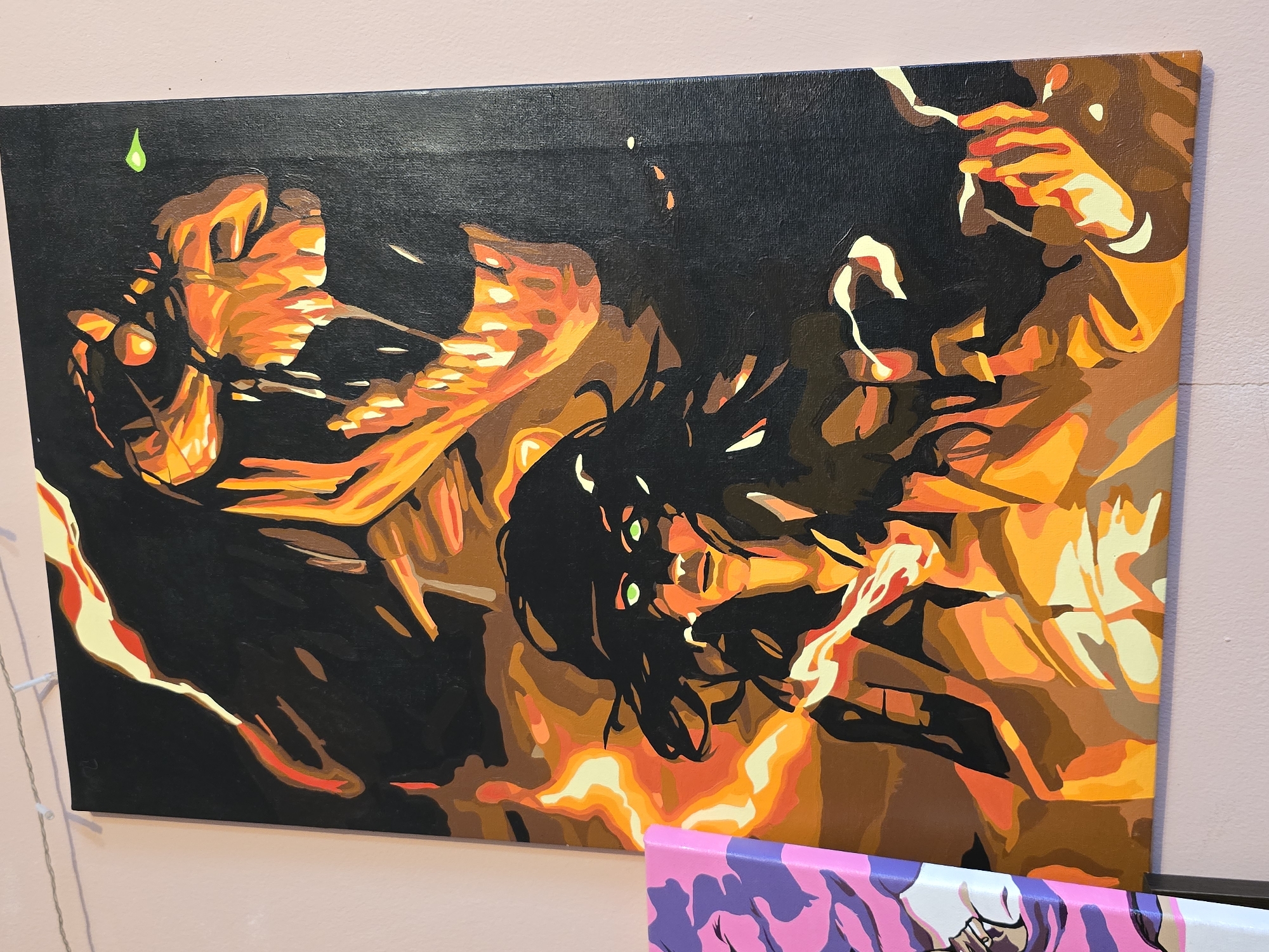A painting on a wall depicts a woman with green eyes and dark hair in front of a fiery dog-like creature. The woman is in the foreground, looking forward with a serious expression, her hands raised and emitting smoke or fire. Behind her, the creature's head is visible, mouth open in a roar, with sharp teeth and an infernal glow. Flames surround the figures, creating a dramatic and intense scene. The painting is rendered in a stylized, graphic manner with bold colors and distinct outlines. The wall behind the painting is a pale pink. To the left of the painting, a portion of another artwork with pink and purple colors is visible. The scene is indoors, likely in a home or gallery setting, though the specific location is unknown.