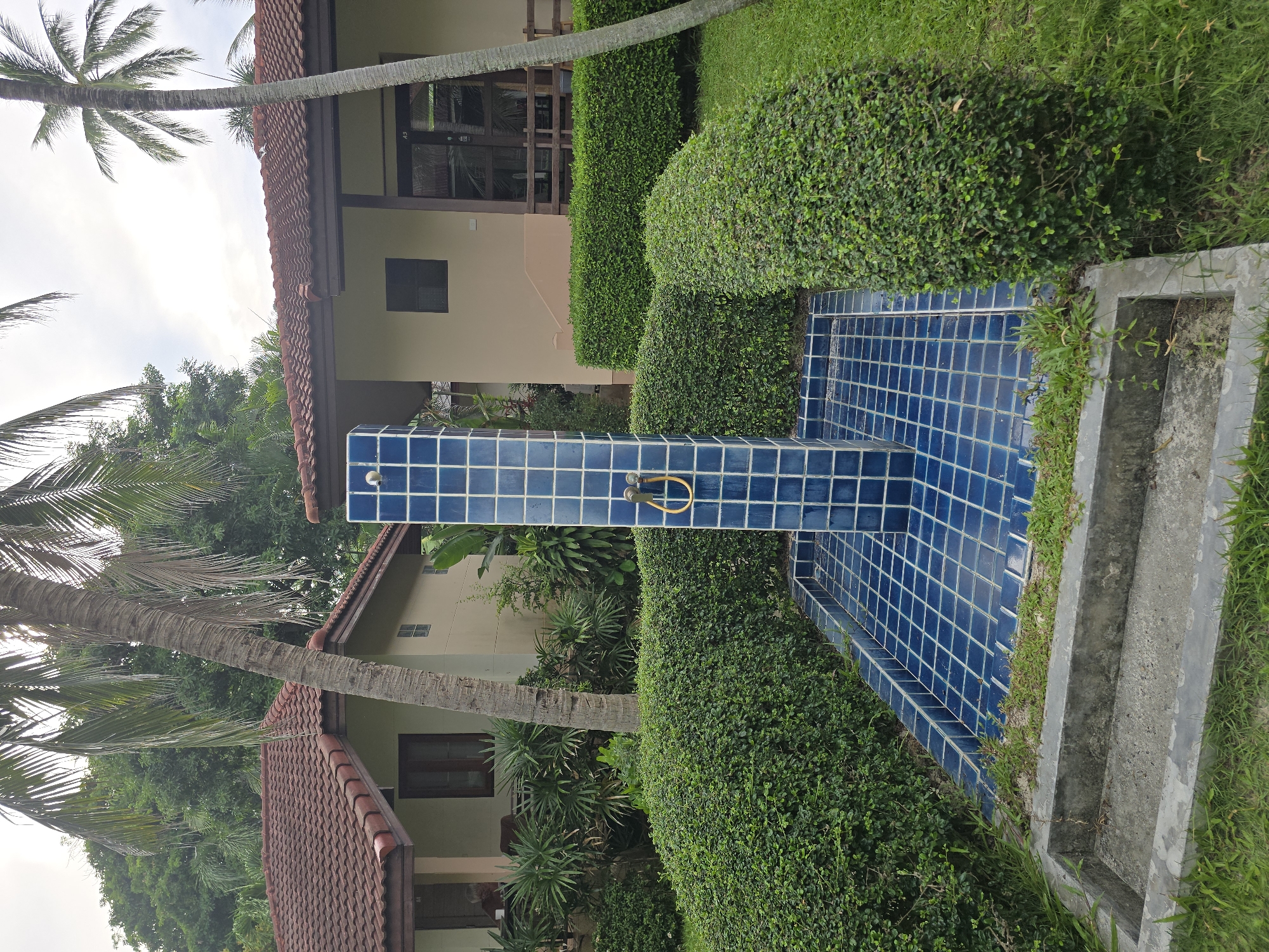 An outdoor shower stall, tiled in blue, stands in the center of a manicured garden. The shower stall is square and features a yellow hose coiled on the side. It is surrounded by neatly trimmed green bushes. In the background, two beige buildings with reddish-brown tiled roofs are visible, partially obscured by lush green foliage and palm trees. The sky is overcast. The scene is calm and appears to be part of a resort or a residential property.
