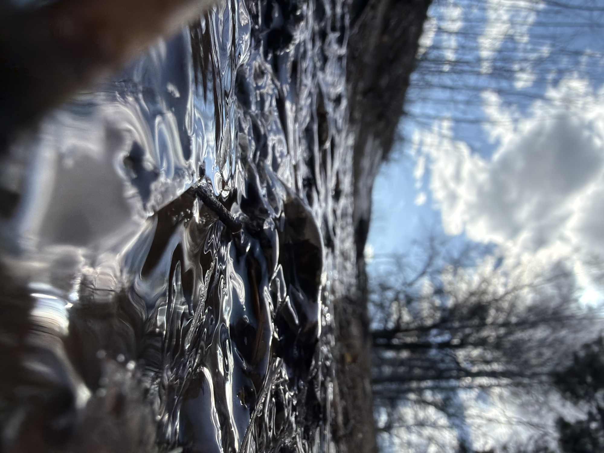 This extreme close-up, low-angle shot presents a dark, amorphous object in the foreground, possibly a distorted reflection on a textured surface like ice or resin. In the background, a blurred scene of bare tree branches against a blue sky with fluffy white clouds is visible. The location of Chaikovsky, Russia, is not discernible from the visual elements present. There are no people visible, and no actions are taking place in the frame. The focus is on texture and light distortion.
