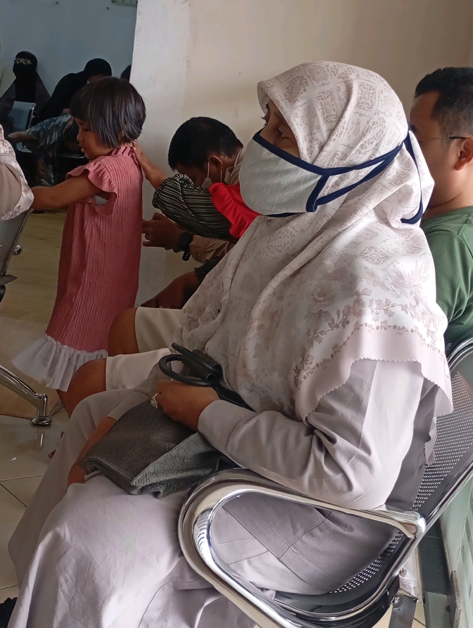 The image captures a scene in a waiting area, possibly a clinic or public facility, in Babelan, Indonesia. In the foreground, a woman wearing a light-colored hijab with a floral pattern and a light blue face mask sits in a metal chair. She is holding a grey handbag on her lap, and a black object, possibly a medical instrument, is resting on the handbag. Her left hand, adorned with a ring, is also visible. The woman appears to be looking downwards.
Behind her, to the right, a person wearing a green shirt is partially visible, with their head turned away from the camera. To the left of the woman in the foreground, a young child in a pink ruffled dress stands, with their back to the viewer. Another person, wearing a striped shirt and a red garment around their neck or head, is visible behind the child, leaning forward and touching the child's back. Further in the background, several other individuals are present, some covered in dark clothing, including one wearing a black hijab.
The environment suggests a communal indoor space with neutral-colored walls and a tiled floor. The lighting appears to be artificial, creating a somewhat flat illumination. The presence of multiple people in a waiting posture, coupled with the medical instrument in the foreground, implies a healthcare-related setting.