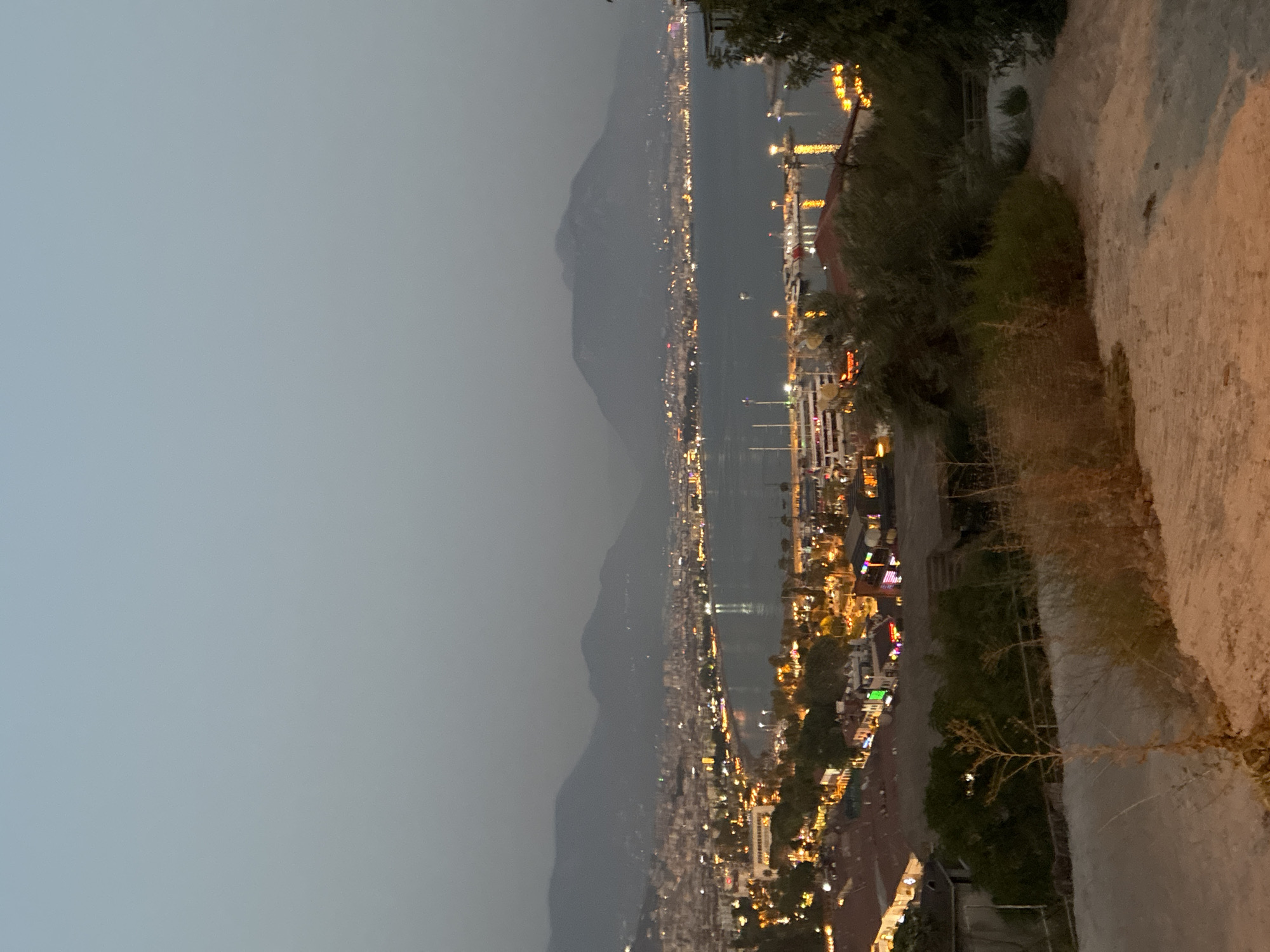 The image shows a panoramic view of Alanya, Turkey at dusk. The city is spread along the coast, with buildings illuminated by lights. A bay filled with boats, including several large passenger vessels, is visible in the middle ground. Mountains form a hazy backdrop to the cityscape. In the foreground, there is a sloped, sandy path with some dry vegetation. The overall impression is that of a coastal city coming alive with lights as night falls.