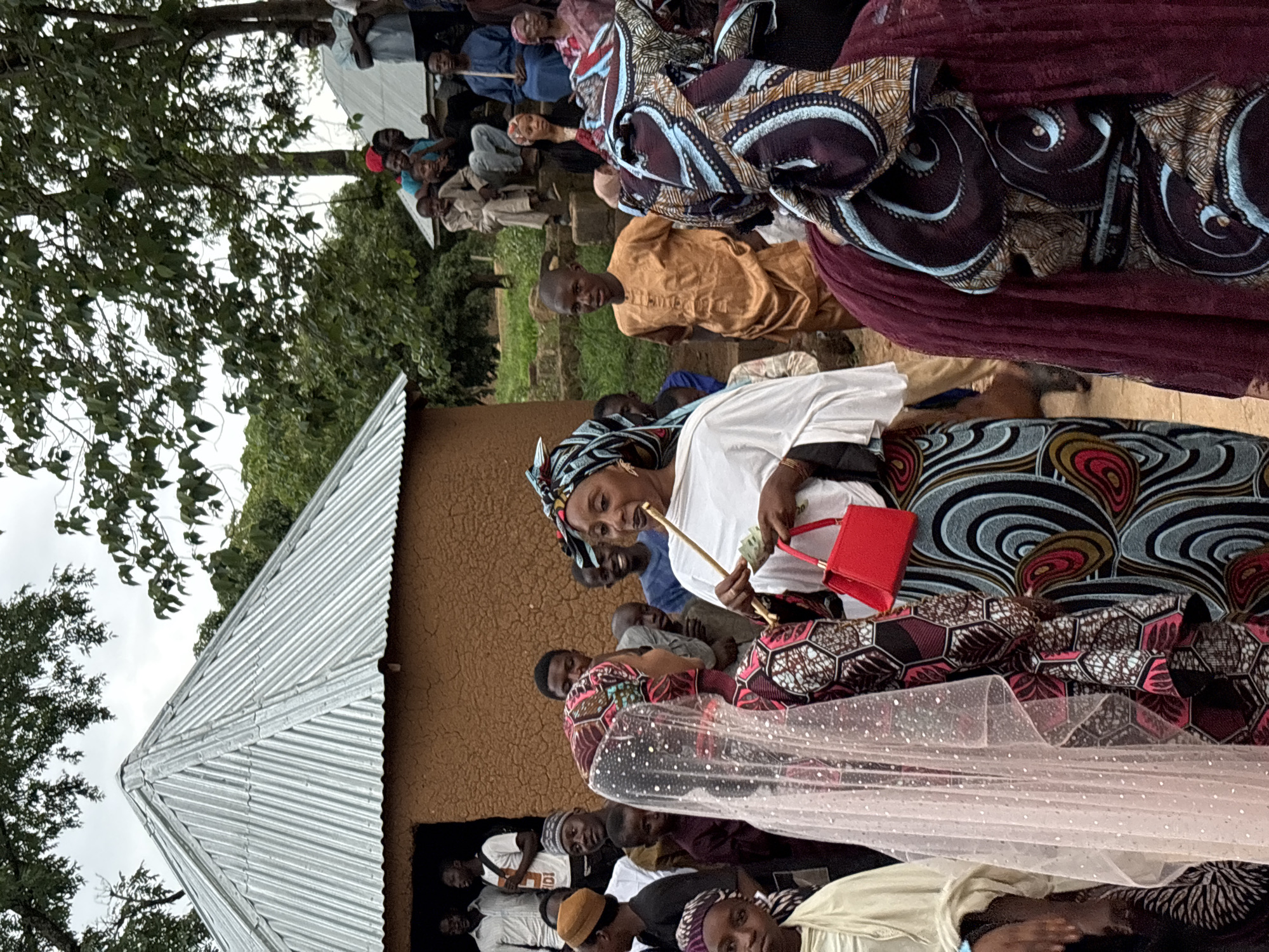 The image depicts a gathering of people outdoors, likely in a rural or semi-urban setting in Jos, Nigeria. In the foreground, a woman wearing a white top and a colorful patterned skirt and headscarf is holding a thin object, possibly a straw or a stick, to her mouth. She is also holding a small red handbag and some money. To her left, another woman is dressed in a patterned outfit and a sheer veil with white dots, suggesting a possible ceremonial or celebratory occasion, perhaps a wedding.
Behind these two women, a crowd of men, women, and children are visible. Some individuals are standing, while others are seated on steps or the ground. The architecture in the background includes a building with a corrugated metal roof and mud walls, typical of some structures in the region. Trees and foliage surround the area, indicating an outdoor environment. The overall atmosphere appears to be one of community engagement and social interaction. The lighting suggests it is daytime, with an overcast sky.