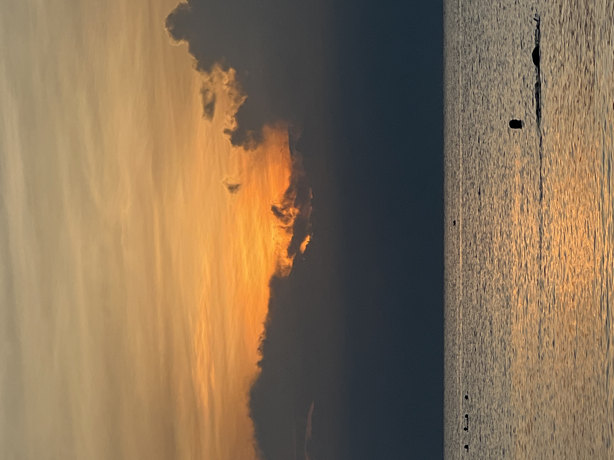 The image captures a serene seascape at dusk, likely in the Ko Pha-ngan District of Thailand. The sky is a canvas of warm, gradient colors, transitioning from a soft yellow-orange near the horizon to a muted orange and then to a darker gray towards the top. Wispy clouds are scattered across the sky, with larger, darker clouds obscuring a portion of the setting sun, allowing glimpses of the intense orange glow behind them.
The ocean stretches out in the foreground, its surface reflecting the warm hues of the sky, creating a shimmering effect. The water is calm, with subtle ripples distorting the reflections. Scattered across the water are several small dark shapes that appear to be buoys or markers, suggesting a swimming or protected area. In the mid-ground, to the right, a person is visible swimming; only their head and shoulders break the surface of the water, with what appears to be a body of water and possibly another person or object close by. Further to the left, a line of four dark, silhouetted heads are also visible, suggesting a group of swimmers or individuals wading in the water. The overall atmosphere is peaceful and tranquil, characteristic of a sunset by the sea.