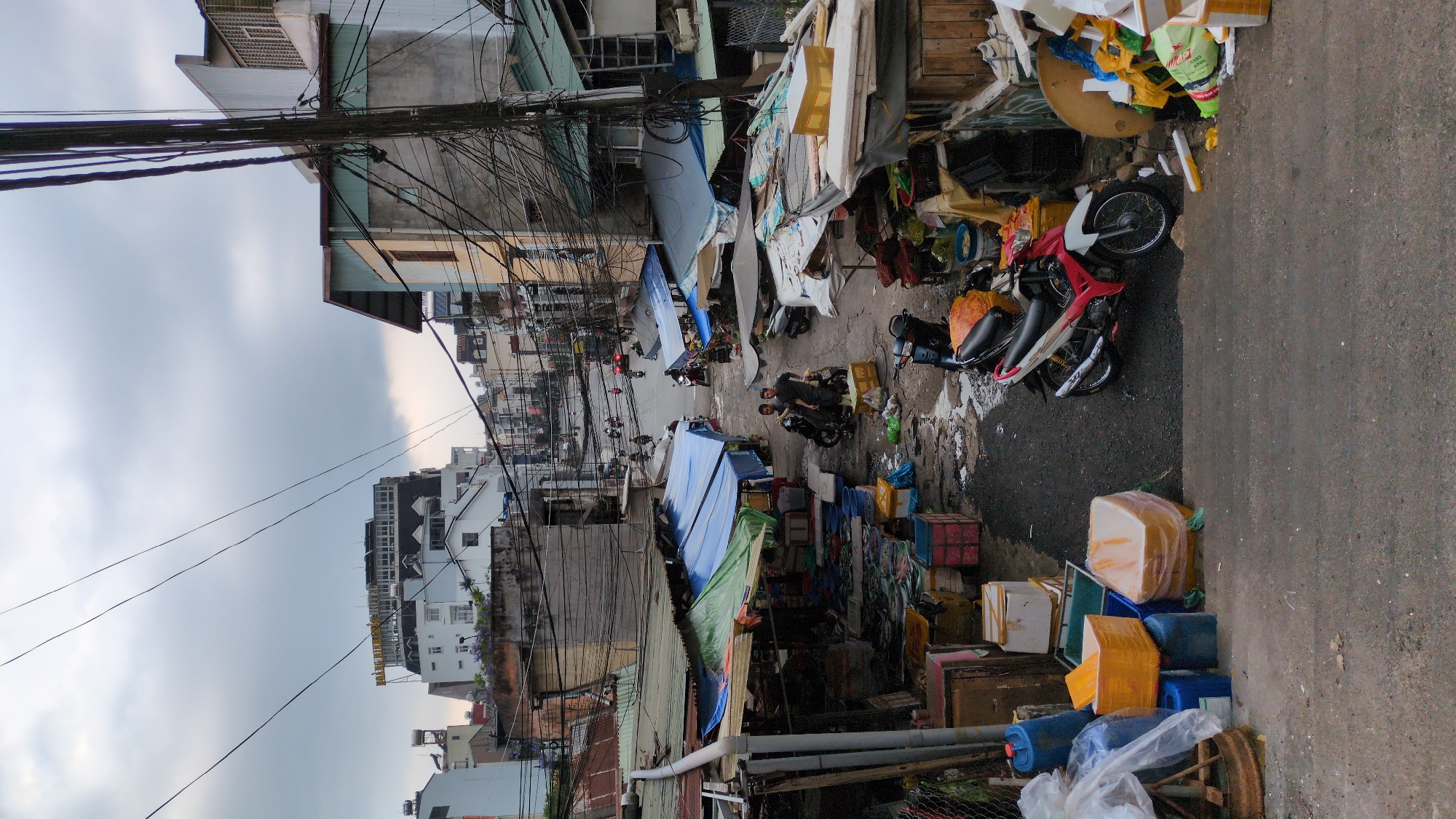 The image depicts a narrow street in Da Lat, Vietnam, densely packed with shops and stalls. The street appears to be a local market area, with various goods for sale, including food and household items, stacked along the sides of the street and under makeshift awnings. There are several motorbikes parked and moving along the street, indicating the primary mode of transportation and business activity. The buildings are a mix of residential and commercial structures, with multiple floors. Overhanging electrical wires contribute to a cluttered urban landscape. The sky is overcast.
