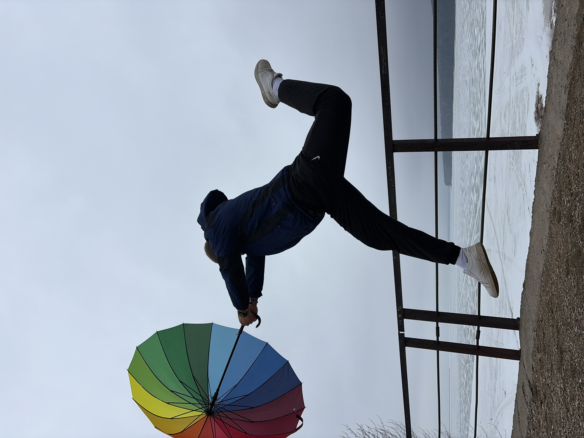 A man is posing upside down with an open rainbow umbrella in his left hand, while holding onto a metal railing. He is wearing a blue jacket, black sweatpants with a Nike logo, white socks, and white sneakers. The background features a body of water, possibly the Kama River, that appears to be partially frozen over, suggesting winter or early spring conditions. The location is the city of Chaikovsky, Russia.
