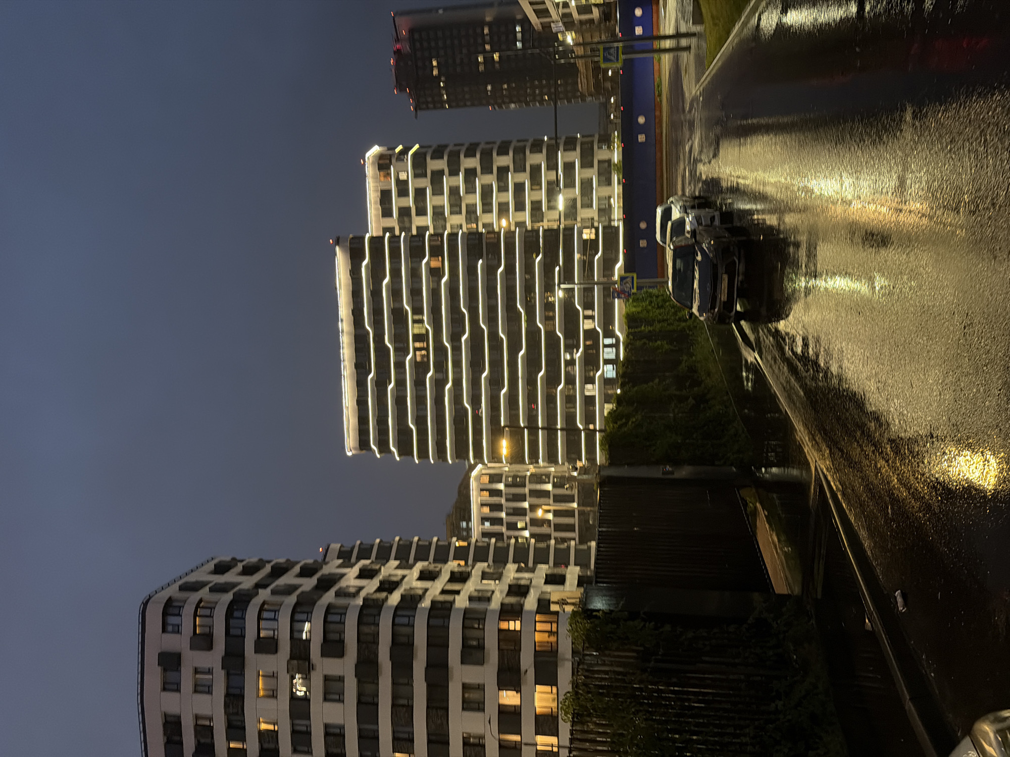 In Moscow, Russia, at dusk, a street scene unfolds with modern architecture and reflections on wet asphalt. Several high-rise buildings dominate the skyline. The building on the left is light-colored with dark horizontal bands and numerous windows, some illuminated with warm light. To its right, another building features a striking design with continuous horizontal white lights outlining its facade, creating a illuminated pattern against the dark sky. Further in the background, a taller, darker building with lit windows is visible, topped with red lights. A row of cars, including a dark SUV in the foreground, are parked or driving on the wet street, their headlights and the building lights reflecting on the slick surface. A dark fence runs along the left side of the street, bordered by green foliage. The sky is a deep, overcast gray, indicating twilight or possibly pre-storm conditions. The overall atmosphere is urban and contemporary, with the wet conditions adding a reflective, almost artistic quality to the scene.