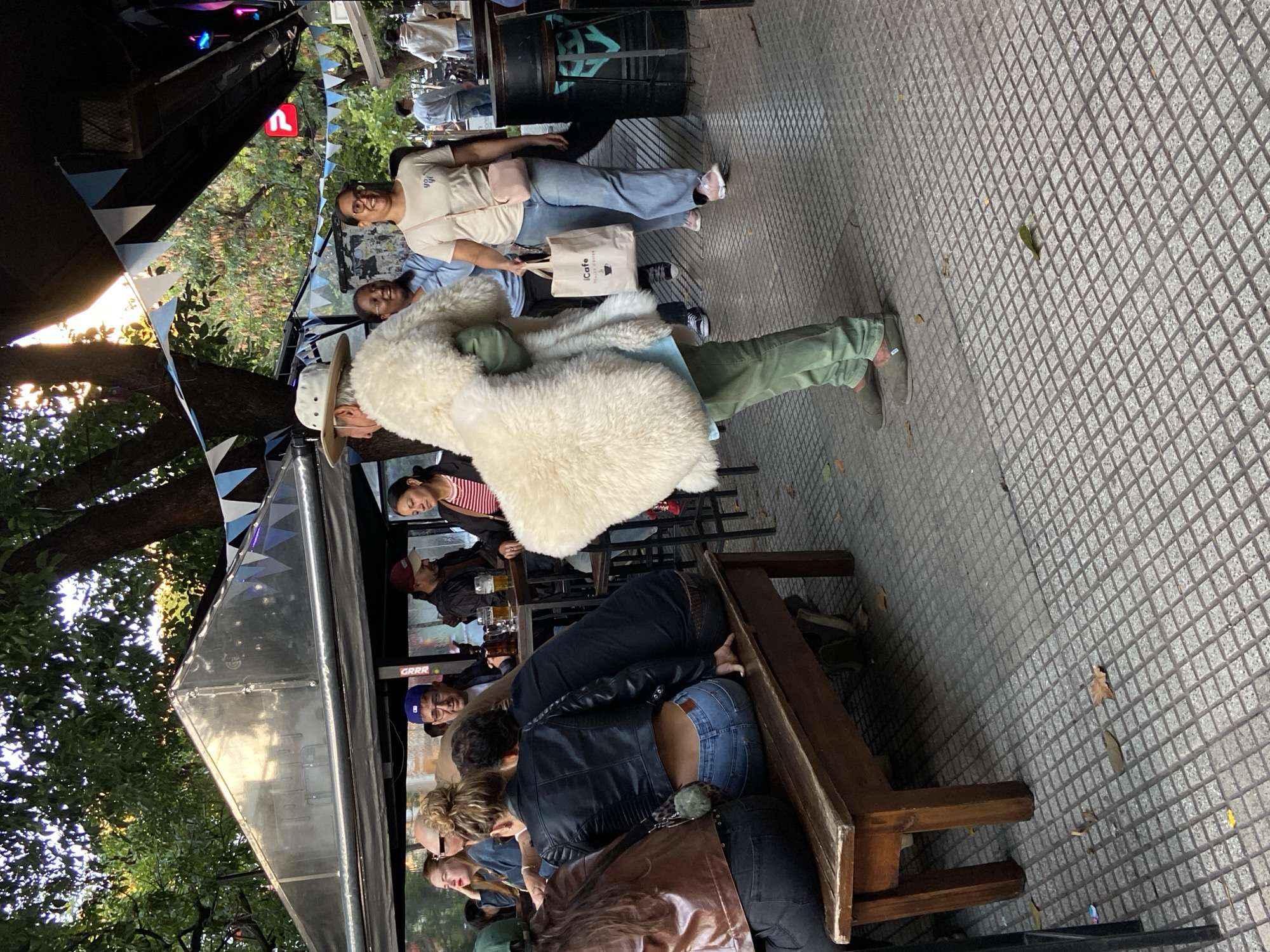 The image shows an outdoor dining area in what appears to be Buenos Aires, Argentina, characterized by tiled sidewalks. People are seated at tables and benches, some drinking. There's a shiny, triangular-shaped food truck with blue and white pennants. One person is wearing a large, fluffy white coat.