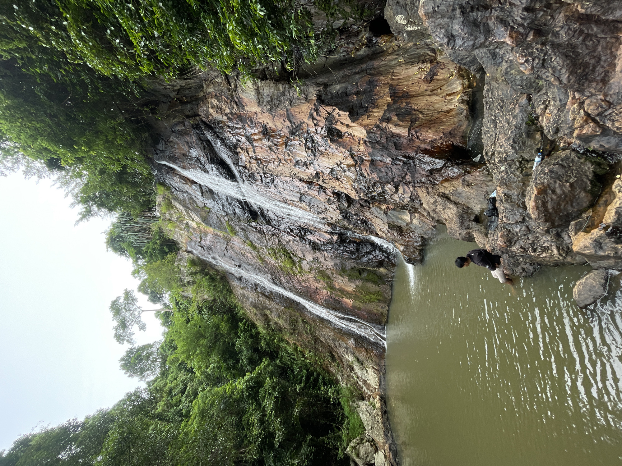 The image shows a waterfall cascading down a rocky cliff face in Baan Thurian, Thailand. The water collects in a tranquil pool at the base of the falls, surrounded by lush green vegetation. A young man is seated on a rock at the edge of the pool, with his feet dangling in the water. He is wearing a black hoodie and light-colored pants. The scene is peaceful and natural, with the sounds of the waterfall likely creating a serene atmosphere. The rocky cliff is wet and glistening, with streaks of mineral deposits adding color to the rock face. Some debris, including bottles, is visible on the rocks near the base of the waterfall. The sky is overcast, casting a soft light over the scene.