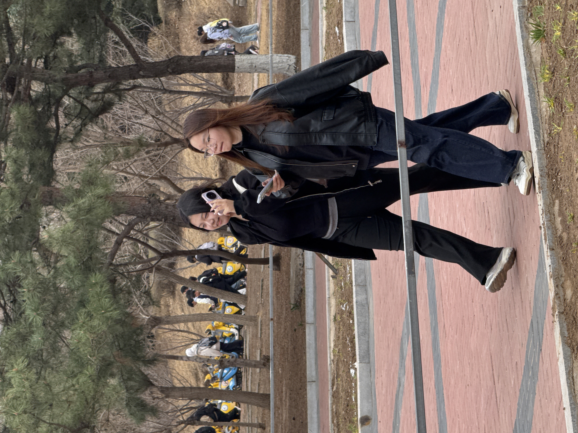 Two women are walking on a brick walkway in what appears to be a park or outdoor area. The woman closer to the camera is taking a photo or video with her smartphone. She has black hair and is wearing a black jacket and matching pants. The other woman is wearing a black leather jacket and dark jeans. Trees and other people are visible in the background, along with a row of parked scooters. The environment suggests a location in a public park or urban green space in Zhongtun, China.