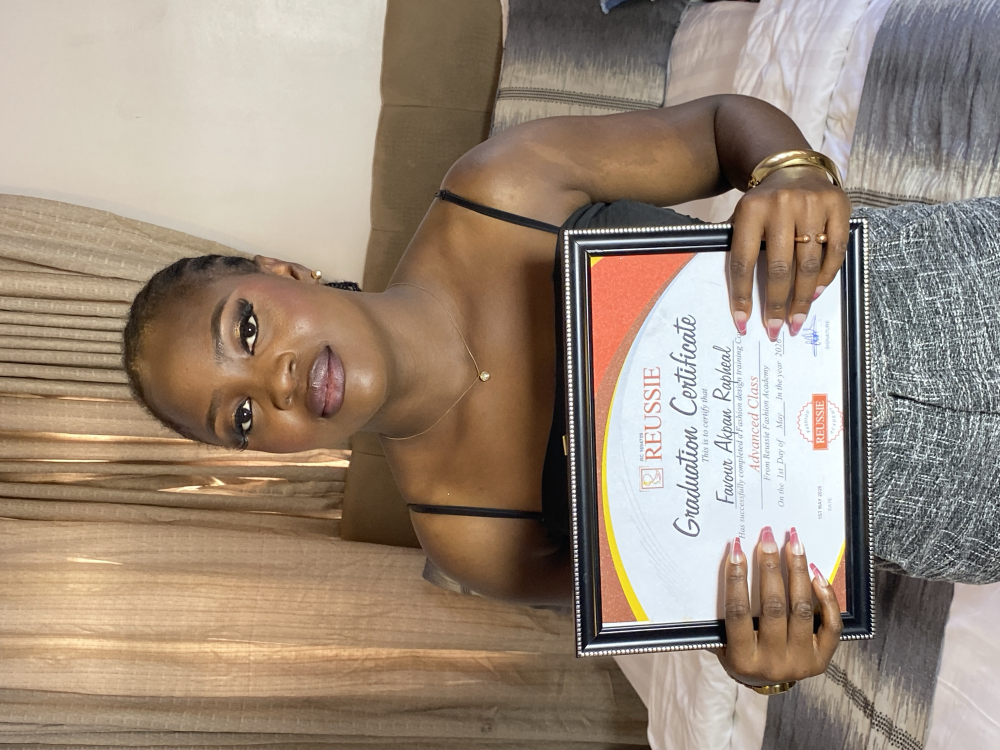 The image, originally rotated 90 degrees counter-clockwise, features a young woman proudly holding a framed graduation certificate in an indoor setting.
The subject is a woman with dark skin and dark braided hair, looking directly at the camera with a calm, confident expression and slightly parted lips. She is wearing a black spaghetti strap top and a grey, textured skirt. Her jewelry includes a delicate gold chain necklace with a small pendant, a thick gold bangle on her right wrist, and a small gold ring. Her long, pointed fingernails display a pinkish ombré design.
The central object is a black-framed "Graduation Certificate." The visible text on the certificate identifies it as from "REUSSIE Fashion Academy" (RC 1694709), awarded to "Favour Akpan Rapheal" for successfully completing an "Advanced Class" in "Fashion design training Course." The certificate is dated "1st Day of May In the year 2026."
The background reveals an interior space, likely a bedroom or living area. A light-colored wall is visible, along with brown textured curtains or fabric on the left. To the right, a bed or sofa with patterned white and grey bedding or cushions can be seen. The lighting is soft, consistent with an indoor environment. The image conveys a sense of achievement and pride.