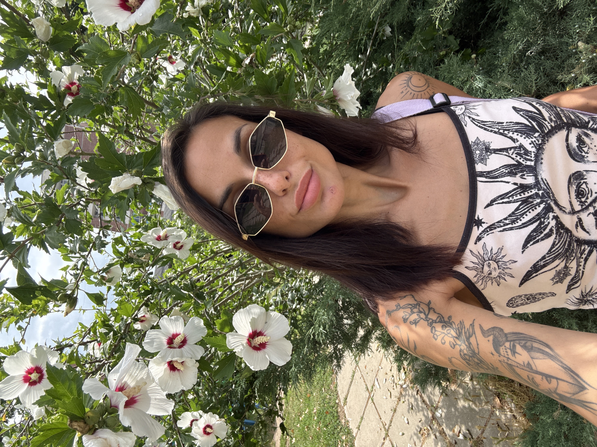 A woman with long, dark hair and sunglasses is standing outdoors in front of a flowering bush. She is wearing a white tank top with sun and moon motifs. Her left arm is visible and has several tattoos. The bush has large white flowers with red centers. The background shows green foliage, a stone pathway, and a building in the distance. This scene is located in the city of Belene, Bulgaria.