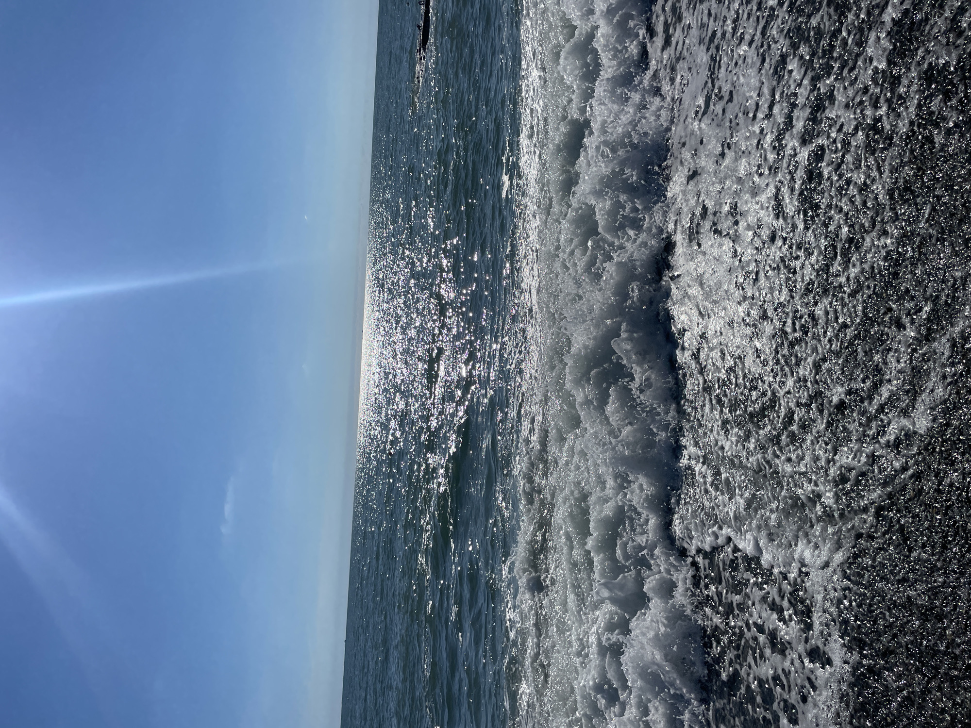 The image depicts a dynamic scene of waves crashing on a dark-colored pebble beach under a clear blue sky. The sun glints brightly off the surface of the water, creating a shimmering effect that extends to the horizon. The waves are white and foamy, with a powerful surge moving towards the shore. The beach appears to be composed of small, dark pebbles. The horizon is clearly defined, separating the vast expanse of the sea from the sky. There are no people or man-made structures visible in the immediate foreground, though a faint dark object, possibly a breakwater or a distant structure, can be discerned on the right side of the image near the horizon. The overall impression is one of a bright, sunny day at the coast. This scene is typical of the Black Sea coast in Sochi, Russia, particularly in areas with pebble beaches.