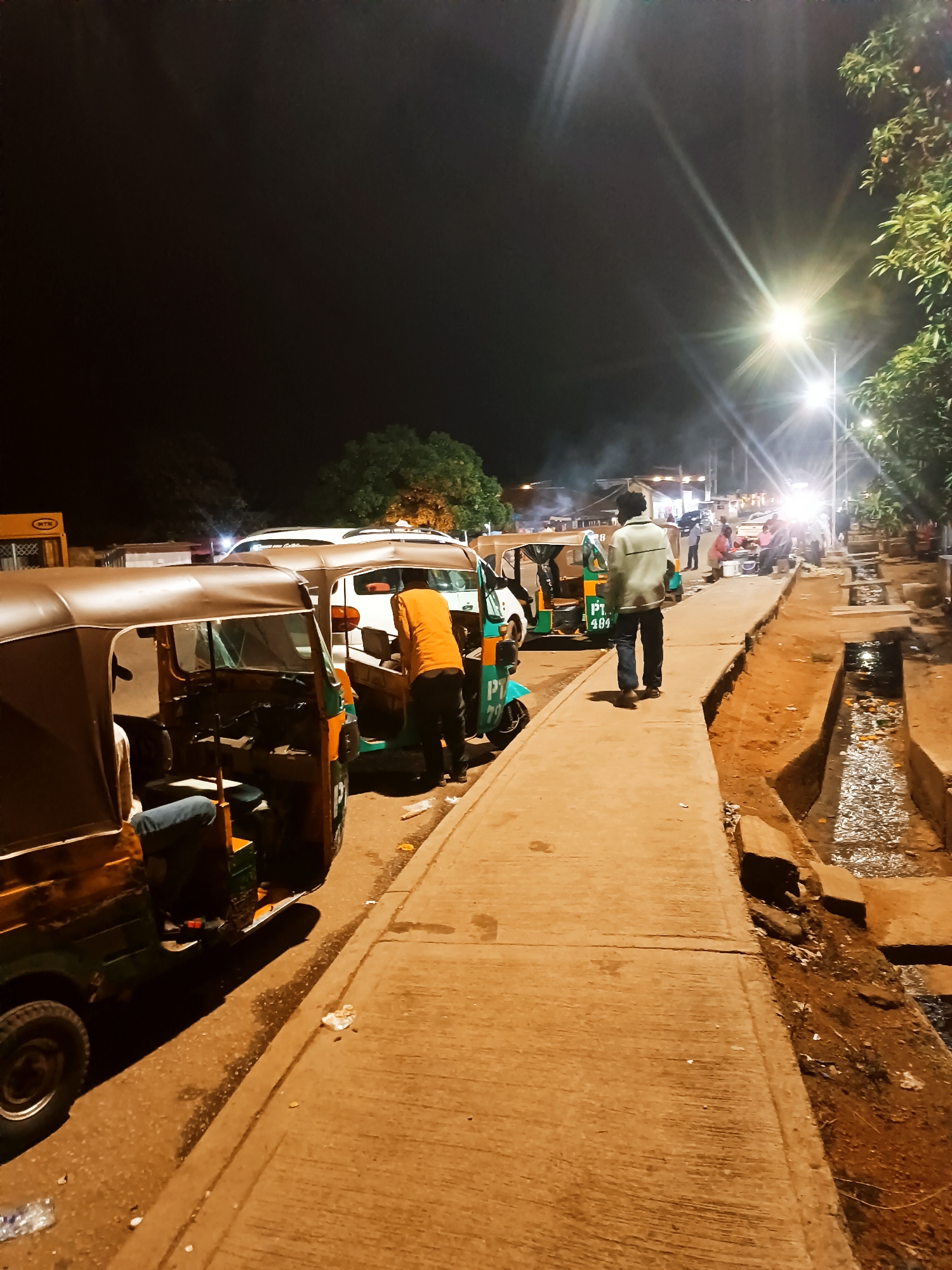 At night in Bukuru, Nigeria, several auto rickshaws are parked along a concrete sidewalk adjacent to a dirt area and a drainage ditch filled with water. A man in an orange shirt is interacting with one of the vehicles. Another man in a light-colored jacket and jeans walks along the sidewalk away from the camera. In the background, there is a gathering of people under bright streetlights, possibly at a food stall or a market area. There is some smoke in the air. The overall lighting is dim, with artificial lights illuminating the scene.