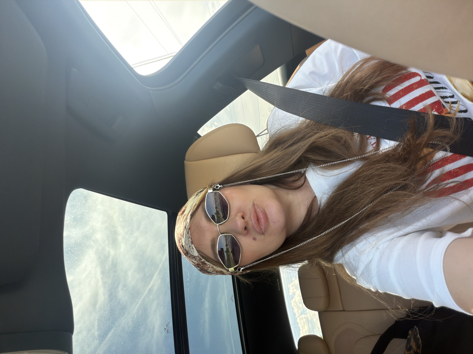 A woman with long brown hair, wearing a headscarf and sunglasses, is sitting in the passenger seat of a car. She is looking at the camera and has her lips pursed. A seatbelt is fastened across her chest. The car has a sunroof, through which the sky is visible. The interior of the car appears to be tan leather. The image was taken in Malinovka, Russia.