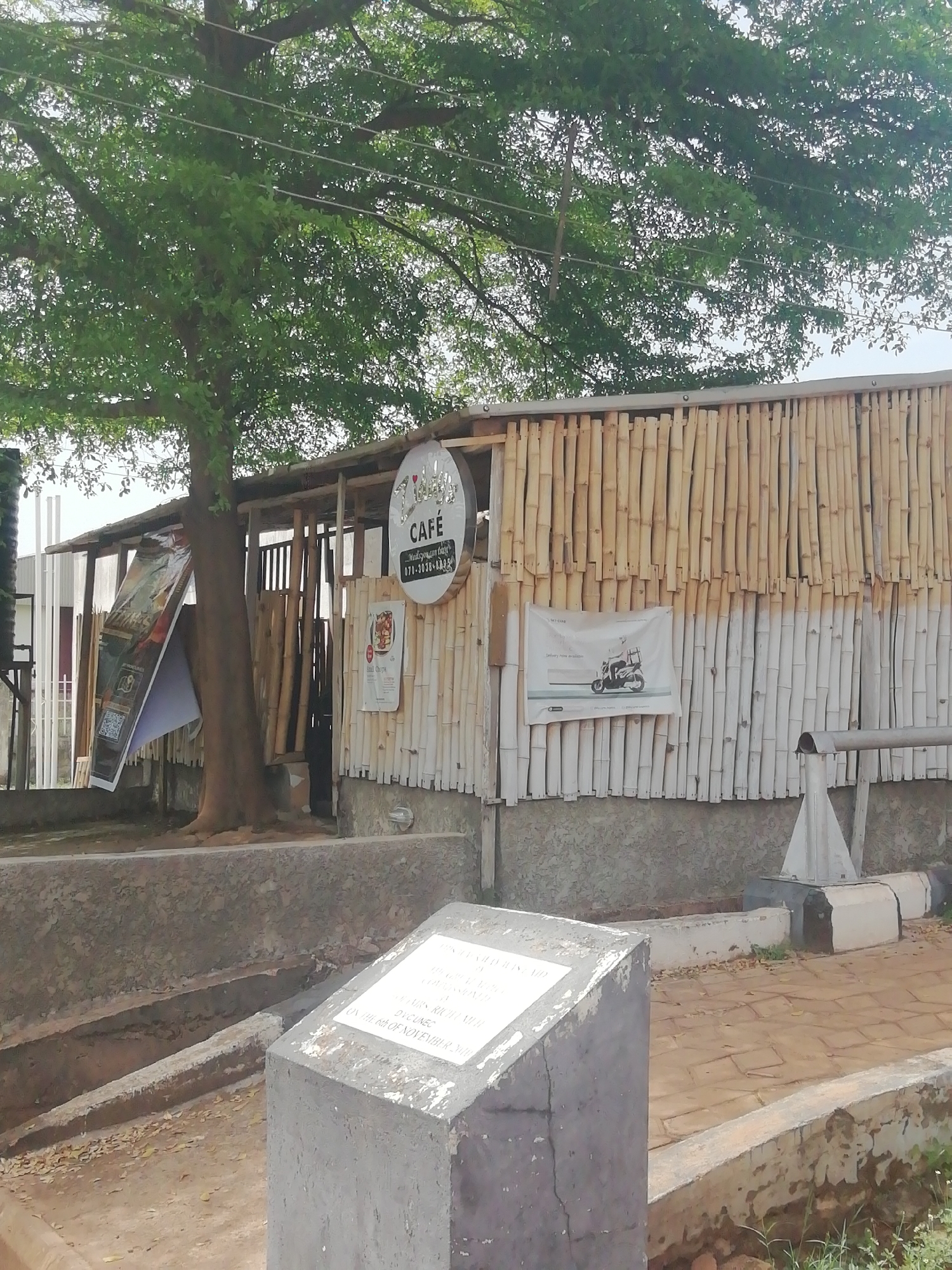 The image shows a small cafe, "7th Heaven Cafe," located in Enugu, Nigeria. The cafe structure is made of bamboo poles, with posters and signs attached to the exterior. A tree is situated in front of the cafe, partially obscuring the view. A stone monument stands in the foreground. The cafe is partially surrounded by a low concrete barrier or planter. The ground is paved with light brown bricks.