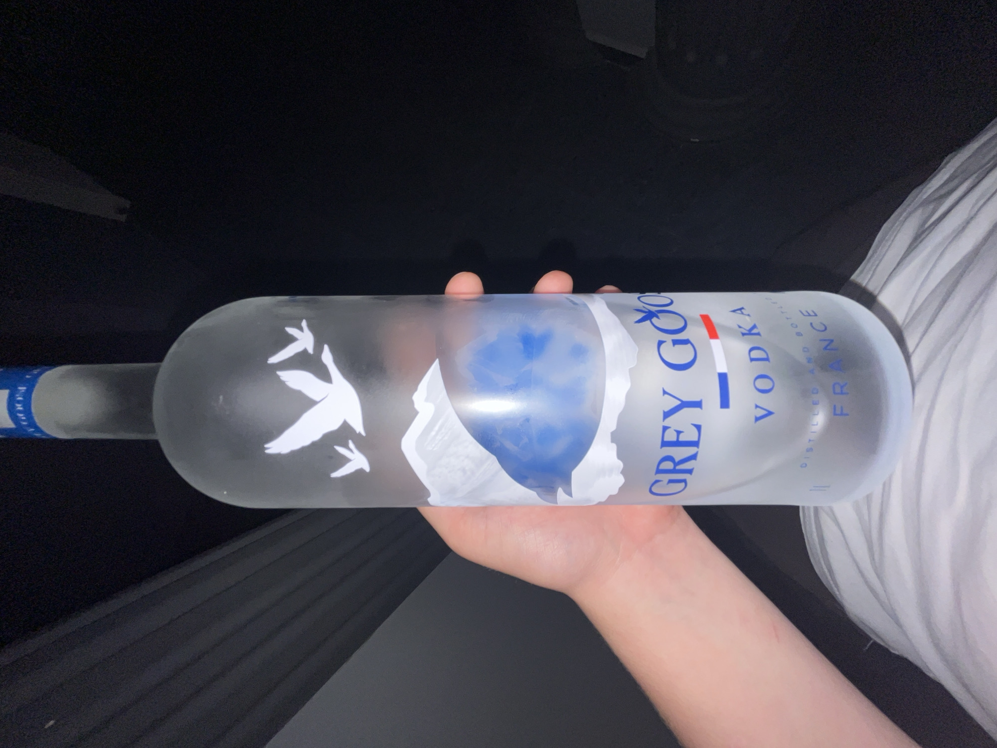 The image displays a hand holding a frosted glass bottle of Grey Goose vodka horizontally in a dimly lit setting. The bottle, primarily clear with a frosted texture, features the iconic white flying goose logo and "GREY GOOSE" prominently printed in blue text, with "VODKA" below it, highlighted by blue and red stripes resembling the French flag. Beneath "VODKA," the text "DISTILLED AND BOTTLED IN FRANCE" is visible. The bottle appears partially full, with a bluish hue visible through the glass.
The hand holding the bottle is light-skinned, with fingers wrapped around the top and bottom of the bottle. The background is predominantly dark, suggesting an indoor environment, possibly at night or in a poorly lit room. To the right, a crumpled white fabric, possibly a sheet or clothing, is visible. To the left and below the bottle, a dark, textured surface, possibly corrugated or ridged, is partially seen. The overall scene conveys a casual, private moment, likely taking place indoors in Peshtera, Bulgaria.