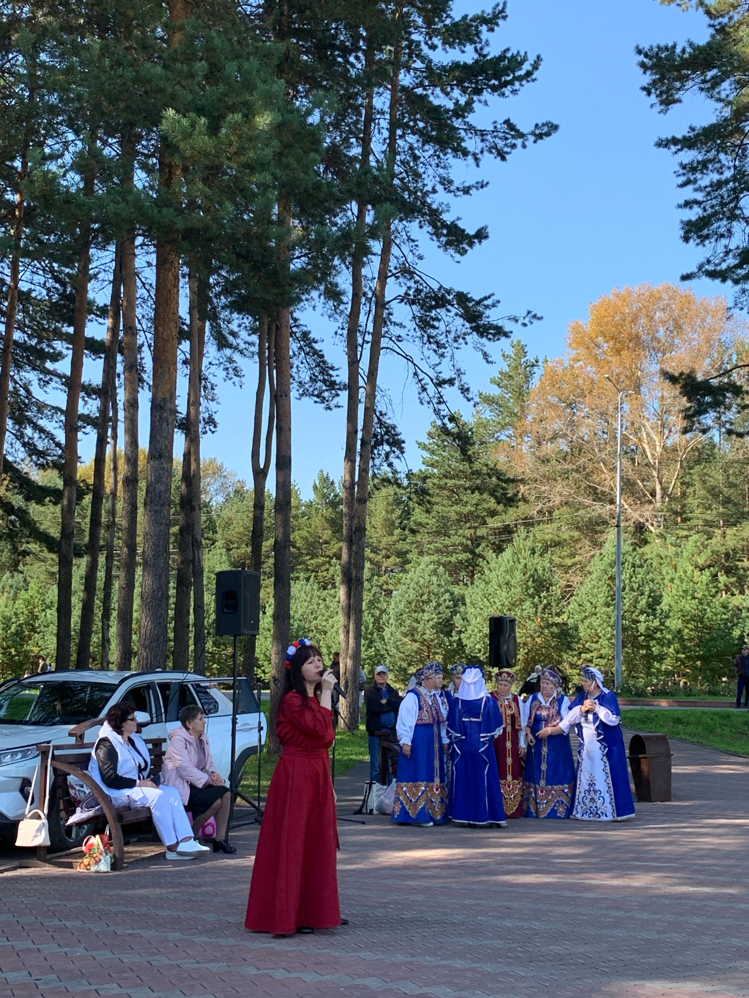 In an outdoor setting with tall pine trees and a clear blue sky, a group of people are gathered on a paved walkway. A woman in a long red dress and a floral headdress is singing into a microphone, standing to the right of the frame. To her left, a group of women in traditional blue and white attire with intricate gold patterns are standing together. A white car is parked behind them. In the foreground, two women are seated on a wooden bench. One woman wears a white vest over black pants, and the other is in a pink jacket. A black speaker is positioned behind the singers, and further to the right, a metal trash bin is visible. The scene appears to be a cultural event or performance in a park-like area within the city of Prokopyevsk, Russia.