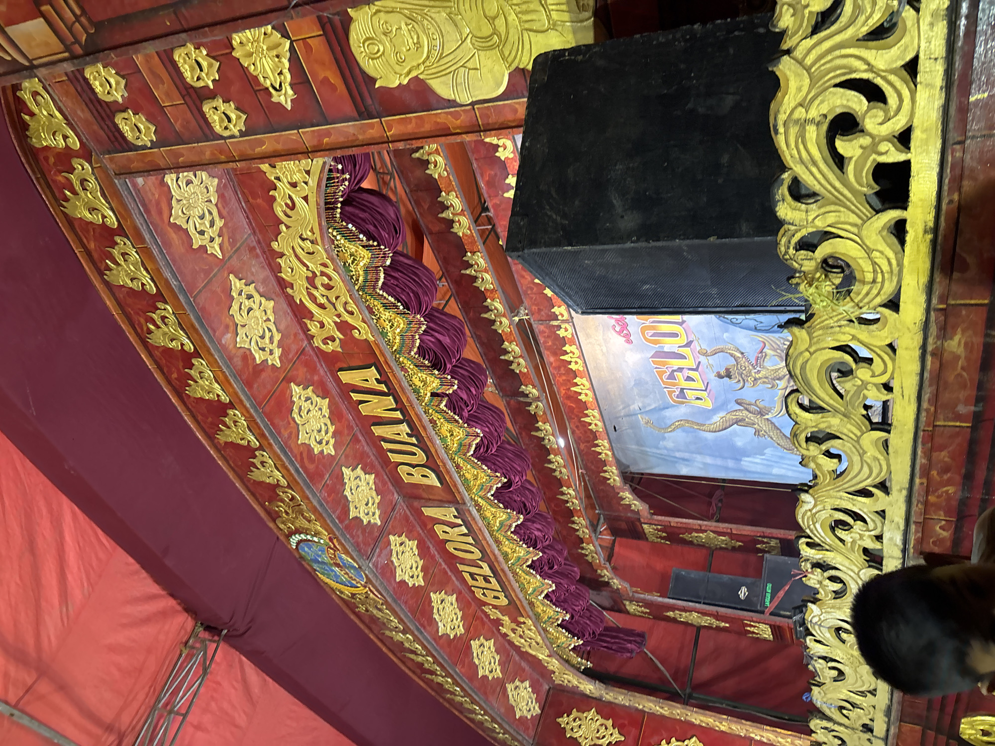 A wide-angle, eye-level, outdoor shot captures a stage set with ornate decorations. The stage features a red backdrop with gold embellishments in floral and brick patterns. A burgundy curtain with gold trim hangs across the upper part of the stage. The words "GELORA BUANA" are painted in yellow letters on the red surface above the curtain. To the right of the stage, a yellow relief carving of a monkey-like figure is visible. A large black speaker box sits to the right of center. Behind the speaker, a blue banner displays the word "GELORA" in large yellow letters, with depictions of dragons on either side. A portion of a person's head, likely a child, is visible in the lower left corner of the frame. The stage appears to be covered by a red tent. This scene is located in Panongan, Indonesia.