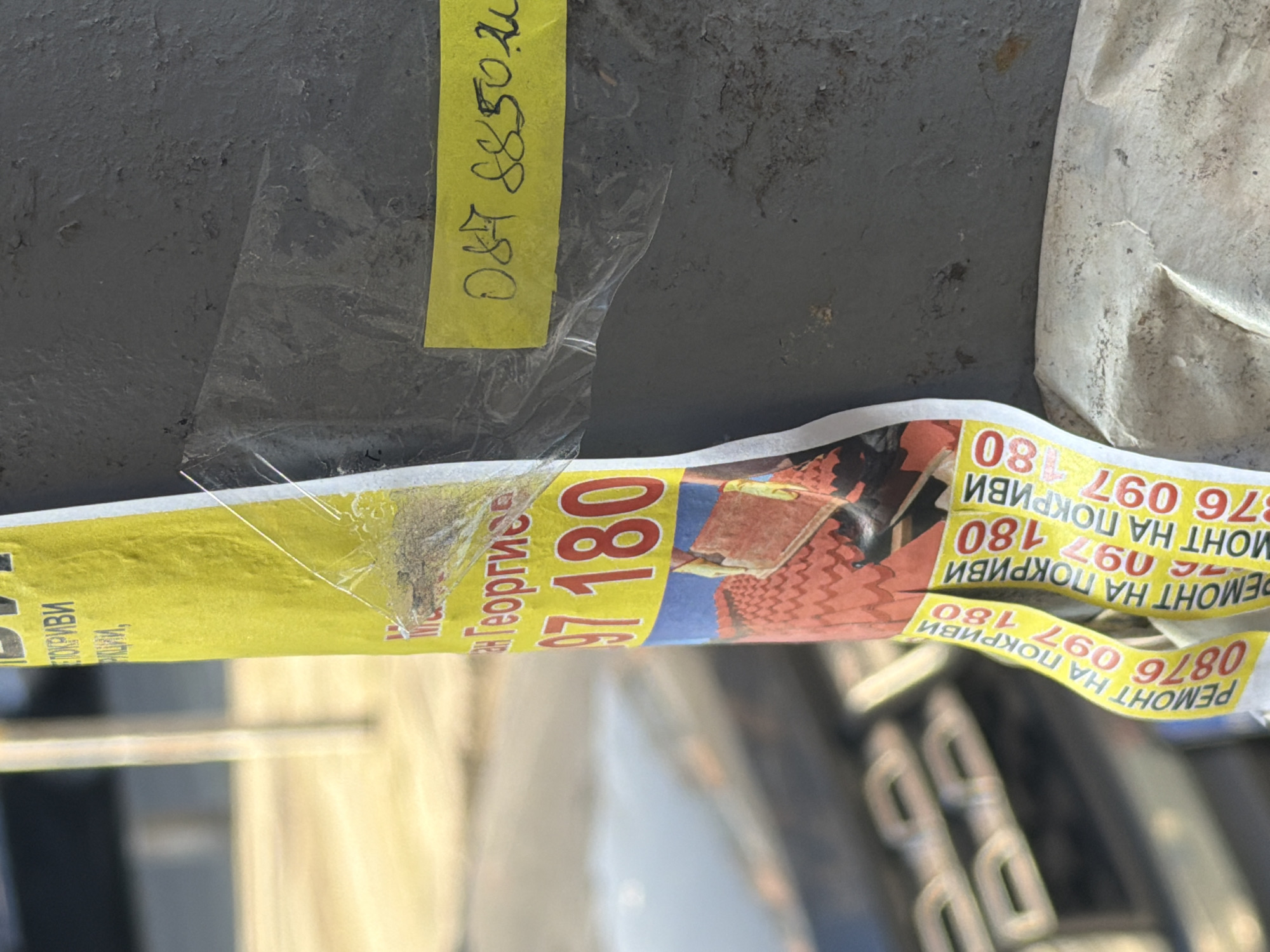 The image displays a close-up, angled view of a textured, dark grey vertical pole or column, against a blurred outdoor background. The primary subject is a partially detached advertisement poster affixed to the pole. The poster is predominantly yellow with red and blue sections, featuring Cyrillic text. Visible text includes "РЕМОНТ НА ПОКРИВИ" (REPAIR OF ROOFS) and several phone numbers, such as "0876 097 180". An illustrative graphic depicting a tiled roof is visible on the lower portion of the poster. The poster is peeling away from the pole, revealing a white backing and some clear adhesive residue or tape, which also secures a smaller, separate yellow strip of paper with a handwritten number, "087 8850 2u", near the top right. The background is out of focus, showing indistinct light-colored vertical elements and a blurry object with stacked, rectangular components. The Cyrillic script on the poster confirms a Bulgarian context, consistent with the city of Burgas; however, the precise location within Burgas is not discernible from the visual information provided. No individuals are depicted.