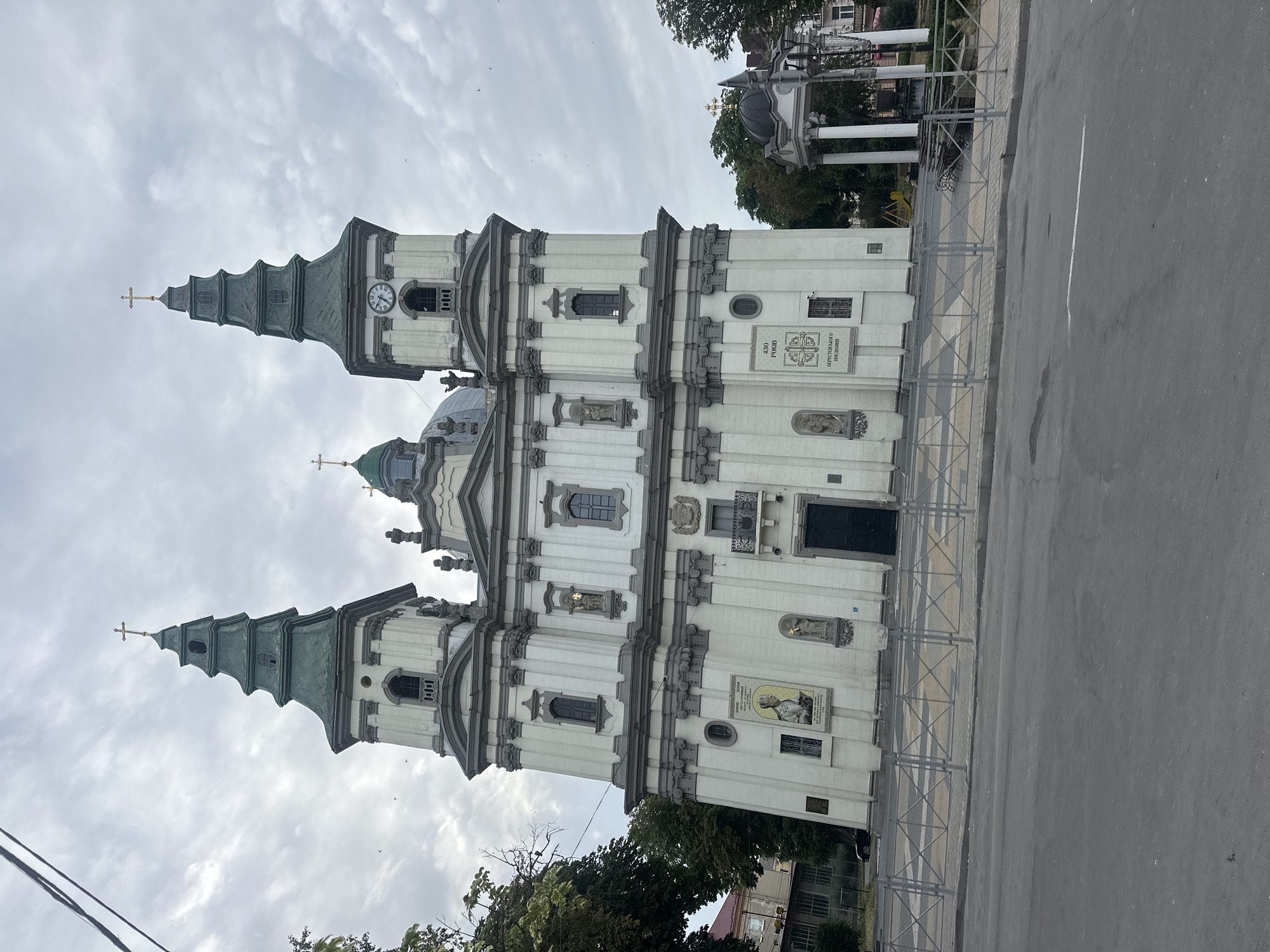 The image shows the Ternopil City Hall, a prominent landmark in Ternopil, Ukraine. The building is a grand, white, Baroque-style structure with two tall towers topped with green spires and crosses. A clock is visible on the right tower. The facade features intricate architectural details, including columns, statues, and decorative moldings. A large banner with an image of a religious figure and text in Ukrainian is displayed on the left side of the building. A second banner with a cross emblem and Ukrainian text is on the right side. The building faces a street, with a grey metal fence separating the sidewalk from the road. Trees and a decorative gazebo with a domed roof are visible to the right of the City Hall. The sky is overcast, and there are a few birds in the sky. The overall scene depicts an urban environment with a focus on the historic architectural significance of the City Hall.