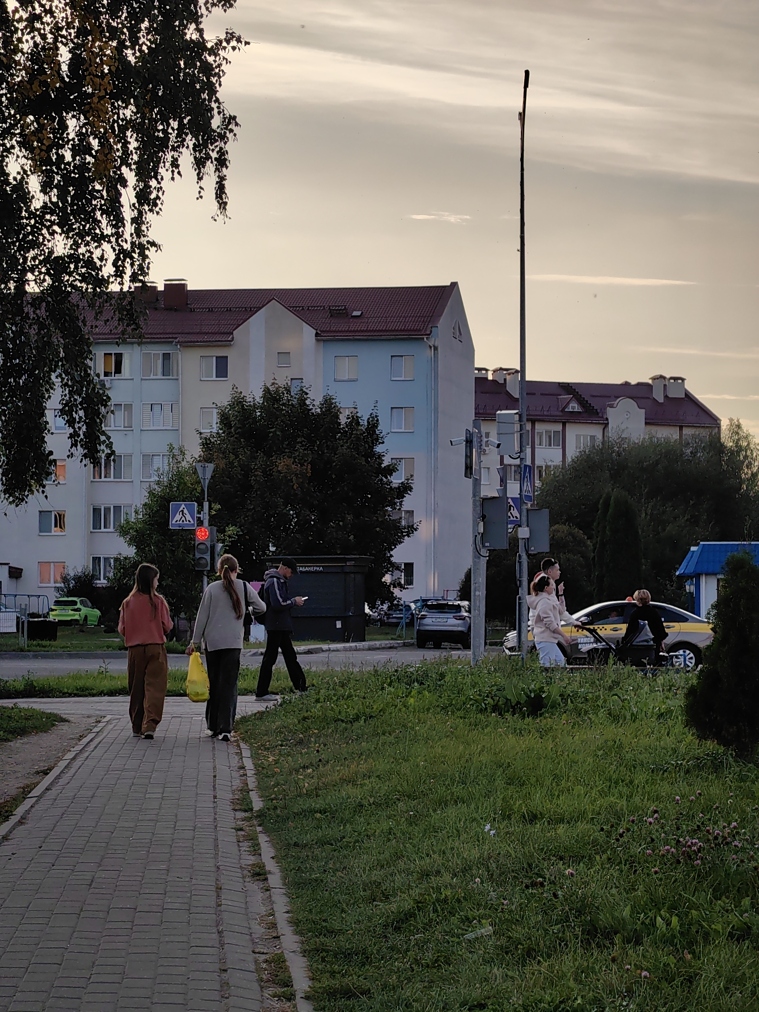 The image depicts a street scene in Fanipal, Belarus. In the foreground, a paved pedestrian walkway runs alongside a grassy area. Two women walk away from the camera on the walkway, one carrying a yellow bag. A man walks behind them. A traffic light displays a red signal, and a pedestrian crossing sign is visible. Beyond the walkway, there is a street with a few parked cars, including a green car. In the background, multi-story apartment buildings with red roofs are visible. A tall lamp post stands to the right of the apartment buildings. Further to the right, a yellow taxi is parked, and several people are standing near it. The sky is overcast with a soft, diffused light, suggesting it is either dawn or dusk.