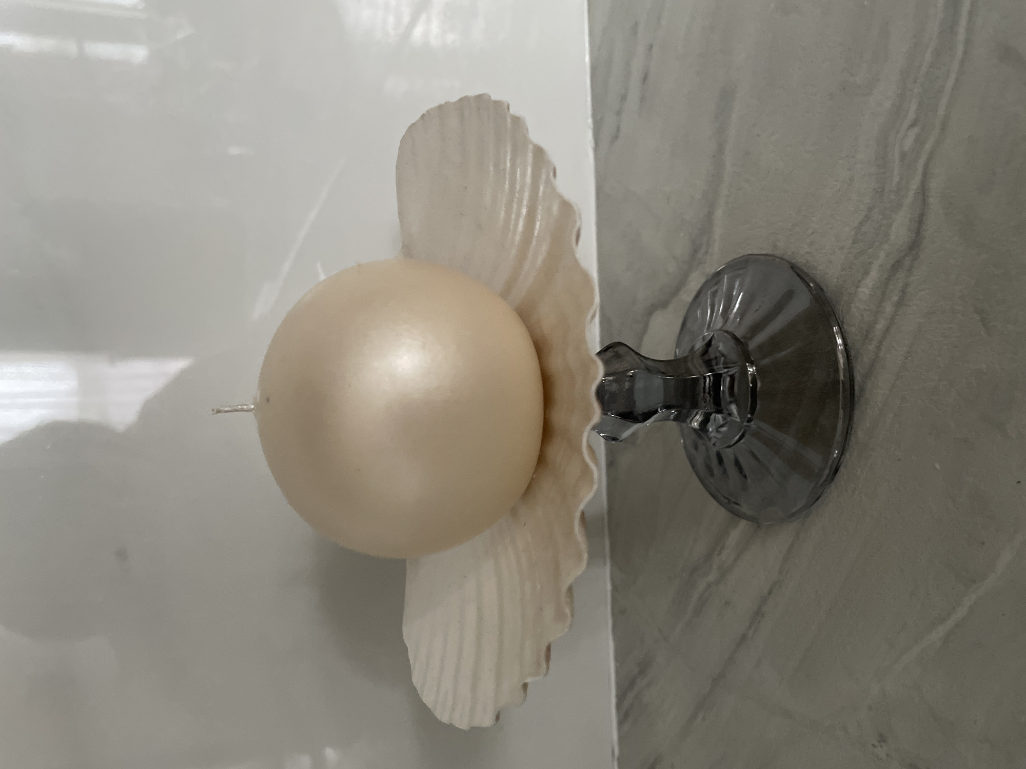 A round, pale yellow candle is mounted on a white seashell, which is affixed to a gray marble wall by a suction cup. The location cannot be determined from the image alone.