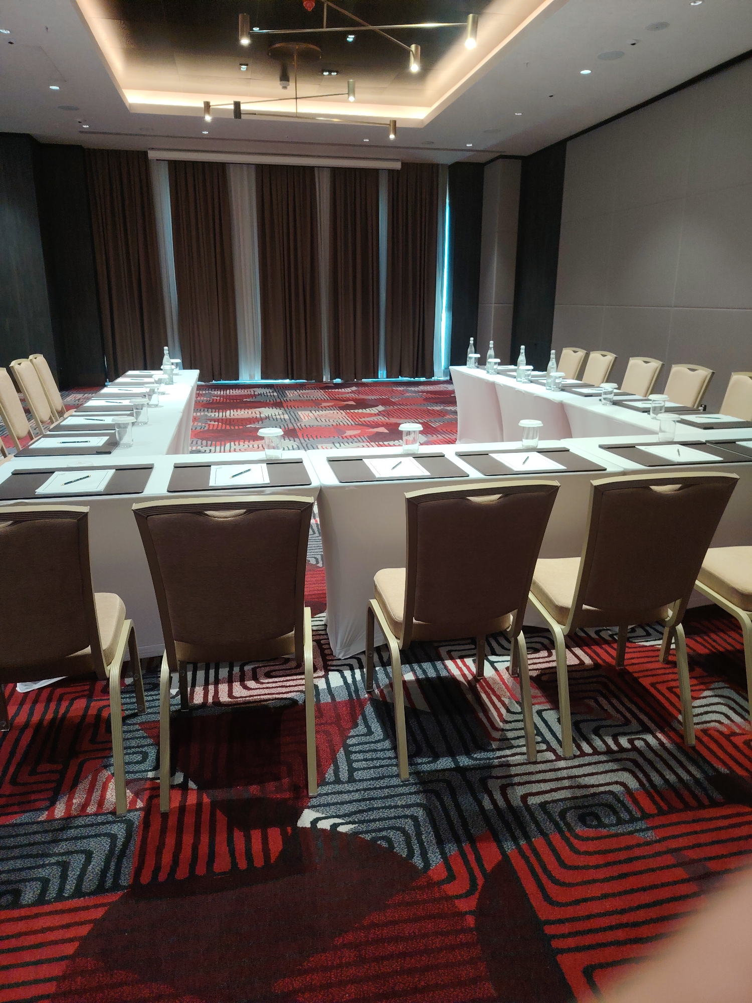 A conference room in Konigil, Uzbekistan, is set up for a meeting with a U-shaped table arrangement. The room features a modern aesthetic with a suspended ceiling that incorporates recessed lighting and a unique light fixture. The walls are paneled in a muted gray on the right, while the left wall appears to be dark. A backdrop of brown and white curtains covers a window or stage area behind the table. The carpet is a striking geometric pattern in red, black, and gray.
The U-shaped table is draped in a white tablecloth, and each place setting includes a dark placemat, a notepad with a pen, and a clear glass. Bottles of water are also strategically placed on the table. Chairs with upholstered brown backs and light-colored seats are arranged around the table, with some chairs facing the camera and others facing away. The lighting in the room is ambient and appears to be coming from the ceiling fixtures. The overall scene suggests a formal business or educational setting, prepared for attendees to participate in a session.