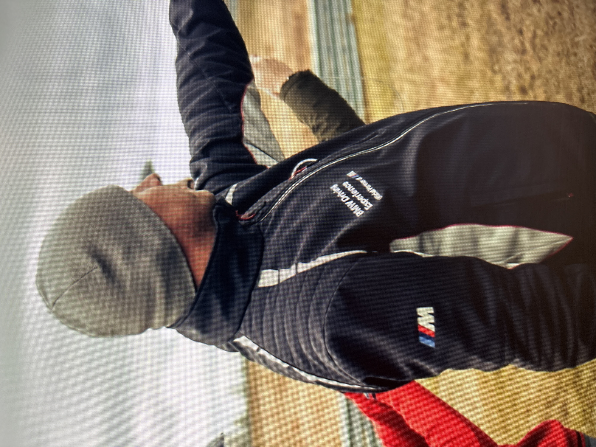 An image shows a man wearing a gray beanie and a black jacket with "BMW Driving Experience" and "Official Partner" text on it, along with the BMW M logo. The man is pointing his right arm towards the right of the frame. He appears to be outdoors, with a blurred background of what looks like a race track and some dry vegetation. Another person wearing a red sleeve is partially visible on the left side of the image, and a hand in a green sleeve is visible on the right. The image does not provide any information about the location being Moscow, Russia.