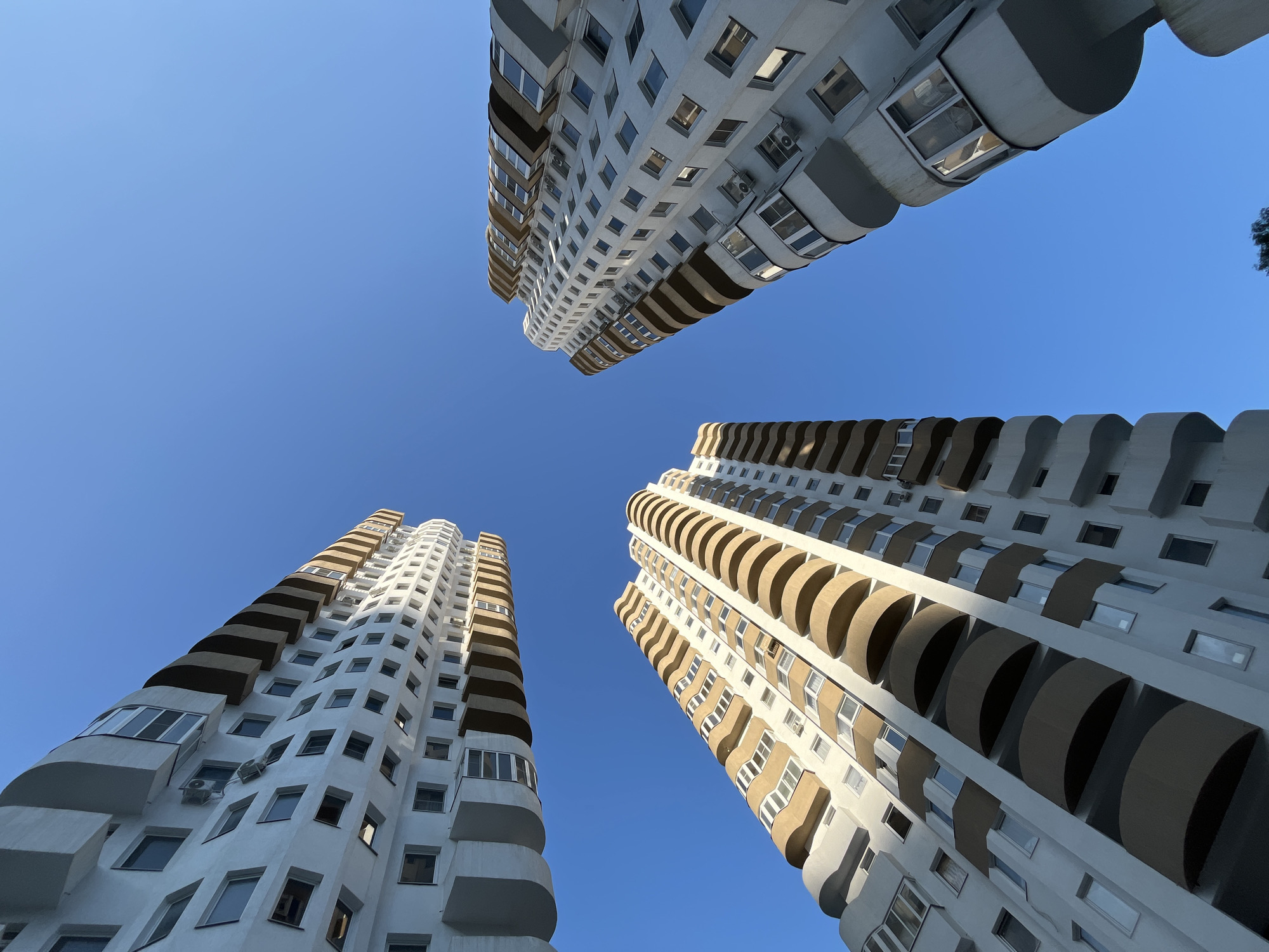 This image shows three tall, modern apartment buildings viewed from a low angle, looking up into a clear blue sky. The buildings are white with beige balconies that are curved and extend outward. Many windows are visible on the facades, along with some air conditioning units. The buildings appear to be part of a residential complex. The scene is an outdoor environment in a city. No people are visible in the image. The overall impression is of a contemporary urban landscape. The buildings are likely located in Sochi, Russia, given the architectural style and the clear, sunny weather, typical of the region.