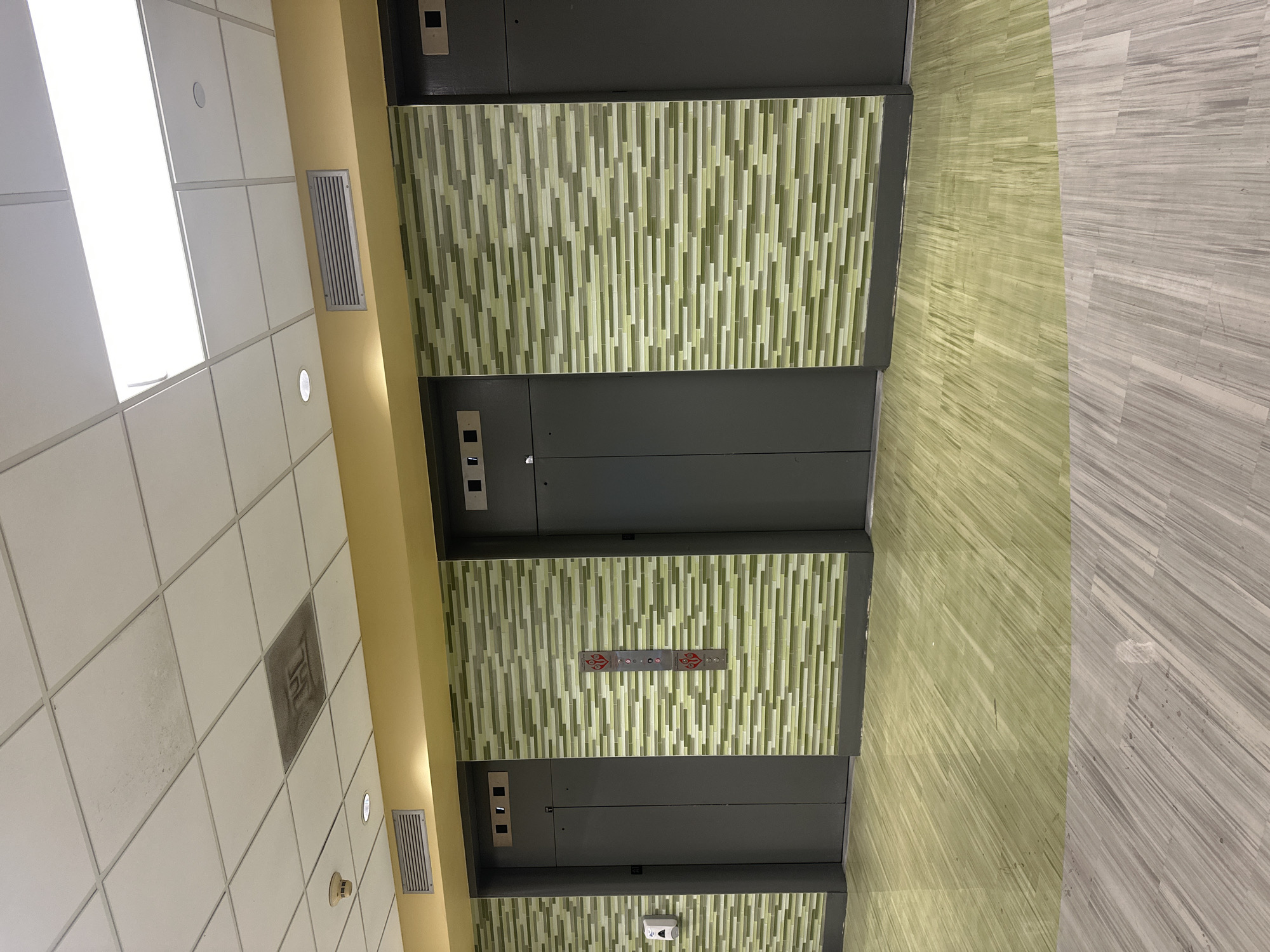 This image, taken in Tampa, United States of America, depicts an indoor commercial or public building hallway or lobby, oriented vertically despite the image's rotation. The focal point is a bank of three elevators with dark grey doors.
Above and below each elevator door, the walls are adorned with decorative mosaic panels composed of vertical rectangular tiles in varying shades of light green, olive green, and off-white. Between the middle and lower elevator doors, a control panel with red decorative flourishes features illuminated arrow indicators and several circular buttons. Similar, simpler buttons are visible on the frames of the upper and middle elevator doors.
To the left of the elevators, a mustard yellow-colored wall extends upwards. This wall features a rectangular metallic air vent. Above, a white ceiling with square acoustic panels is visible, illuminated by both flush circular lights and larger rectangular light panels. A circular smoke detector is mounted on the ceiling. One ceiling tile appears darker and stained, possibly bearing an indistinguishable faded symbol.
The floor is divided into two distinct sections. Closer to the elevators, the floor is a glossy light green, possibly a polished concrete or a type of laminate, displaying a subtle striped texture. Further away to the right, the floor transitions to a lighter grey material, also with a wood-grain or streaked pattern. No people or specific activities are visible in the scene, which appears well-lit and conveys a modern aesthetic.