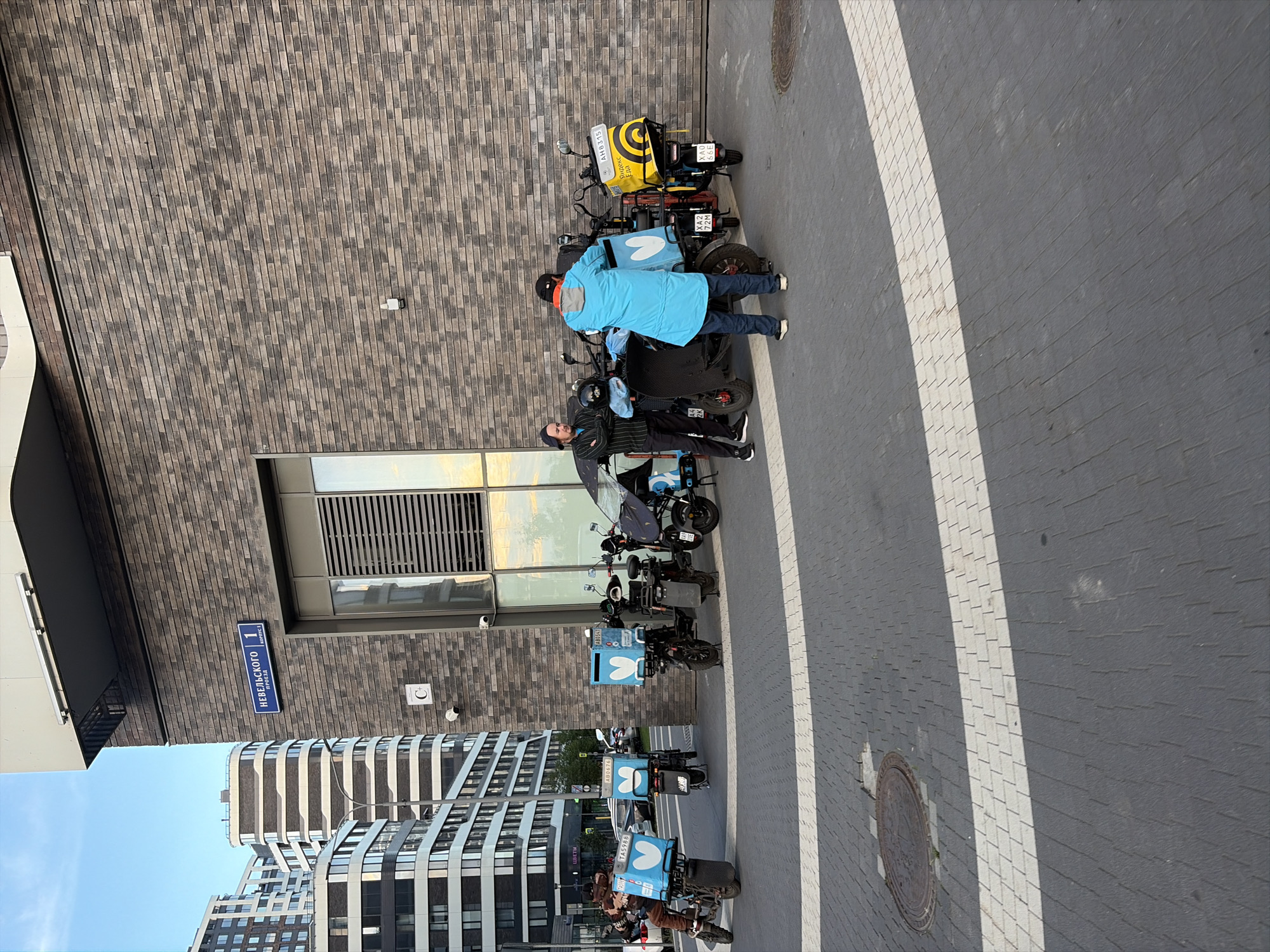 The image depicts a street scene in Moscow, Russia. In the foreground, two men are standing near several delivery scooters parked along a sidewalk. One man, wearing a light blue jacket and jeans, appears to be interacting with a scooter. The other man, dressed in a striped long-sleeved shirt and dark pants, stands nearby. The scooters are equipped with blue delivery boxes, some featuring a white heart logo. One scooter has a yellow delivery box with the Yandex.Eda logo. In the background, modern buildings with distinctive architectural designs are visible. A street sign with Russian text, "НЕВЕЛЬСКОГО 1", is affixed to the brick building wall. The ground is paved with gray and white interlocking bricks, and a manhole cover is present on the sidewalk. The sky is clear and blue.