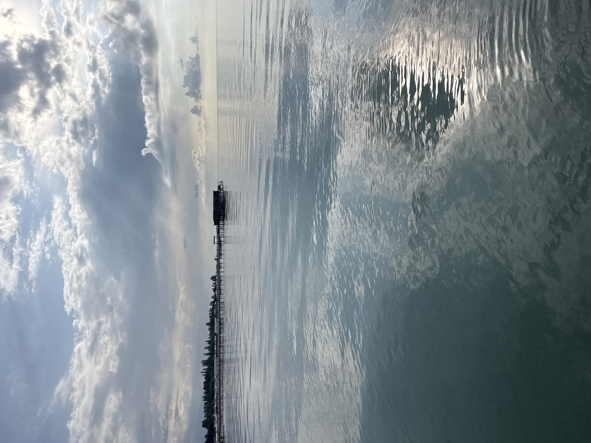The image shows a serene view of a body of water, likely Issyk-Kul Lake, with a pier stretching into the distance in Sary-Oy, Kyrgyzstan. The sky is partly cloudy, with sunlight breaking through in some areas, casting reflections on the water's surface. Along the shoreline, a line of trees can be seen, and a wooden structure, possibly a boathouse or small building, stands on stilts over the water. The water itself is calm, with gentle ripples. There are no people visible in the scene.