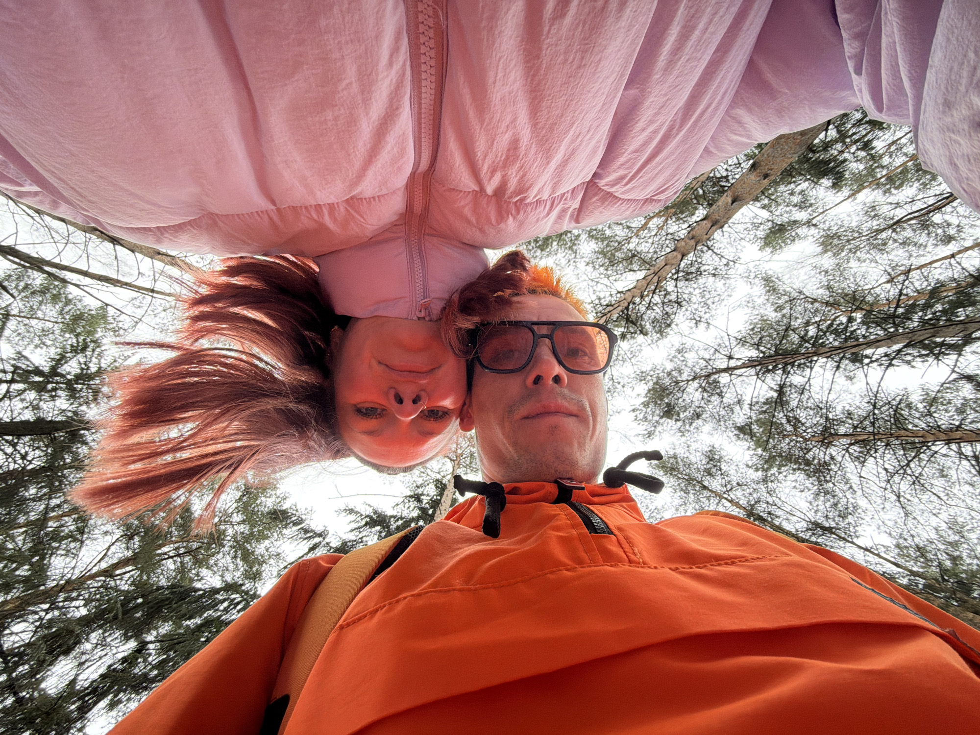 The image presents a unique low-angle selfie featuring two individuals, a female and a male, looking down directly into the camera lens. The perspective makes them appear upside down, or as if the camera is placed on the ground, looking up at them.
The female is positioned in the upper part of the frame, her head angled downwards. She has long, reddish-brown hair, which appears to flow towards the bottom of the image due to the angle, and is wearing a light pink puffer jacket that fills much of the top half of the photo. Her expression is neutral with a slight, subtle smile.
Below her, closer to the viewer, is a male figure. He has short, reddish-blonde hair and is wearing dark-rimmed glasses. He is dressed in a vibrant orange anorak or jacket, and a brown backpack strap is visible over his left shoulder. His gaze is also directed downwards at the camera with a neutral expression.
The background consists of a clear, bright, yet overcast sky, suggesting a cloudy daytime. Numerous tree branches, both bare and some with evergreen needles (likely pines), are visible against the sky, indicating an outdoor setting within a forest or wooded area. The general atmosphere suggests a temperate climate, possibly late winter or early spring in Chaikovsky, Russia. No visible text can be discerned on any of the subjects' clothing or objects. The image is a playful, close-up portrait in a natural environment.