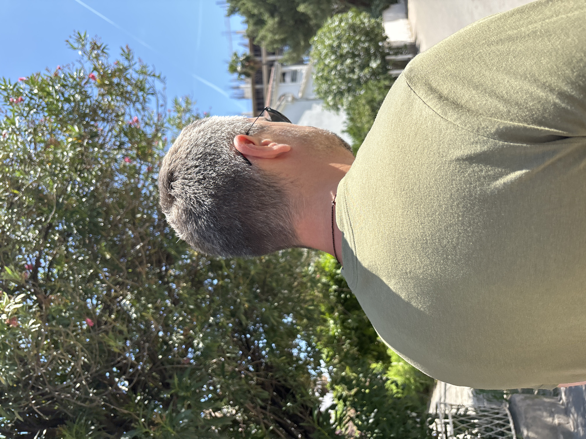 The photograph shows a person from the side, looking up. They have short, gray hair, a beard, and are wearing sunglasses perched on their head. The person is wearing a light green t-shirt. In the background is a tree with pink flowers, and a building under construction. The image appears to be taken in the city of Budva, Montenegro. A silver metal grid fence is also visible.