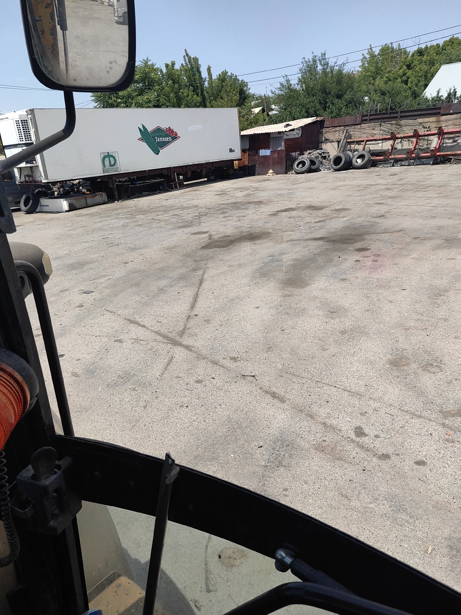 The image depicts an outdoor industrial or storage area on a clear, sunny day, likely in Shymkent, Kazakhstan. In the foreground, partially visible from the perspective of a vehicle's interior, is a black frame with an orange cylindrical object to the left, possibly a hose or conduit. A metal lever or control is also visible. The middle ground is dominated by a white refrigerated trailer truck with a blue and green logo featuring a green bean and red berries, identified as "Janssen." To the left of the trailer, a portion of a white refrigerated unit is visible, with the word "KRONE" printed on it. Behind the truck, a small, weathered, dilapidated structure with a corrugated metal roof stands adjacent to a rusty metal fence. A pile of used tires is visible in front of this structure, along with additional tires scattered in the area. Further in the background, red metal racks are stacked, and lush green trees line the perimeter of the scene. The ground is paved with asphalt or concrete and shows signs of wear and discoloration, including oil stains and tire marks. The sky is a clear, light blue. There are no people visible in the image.