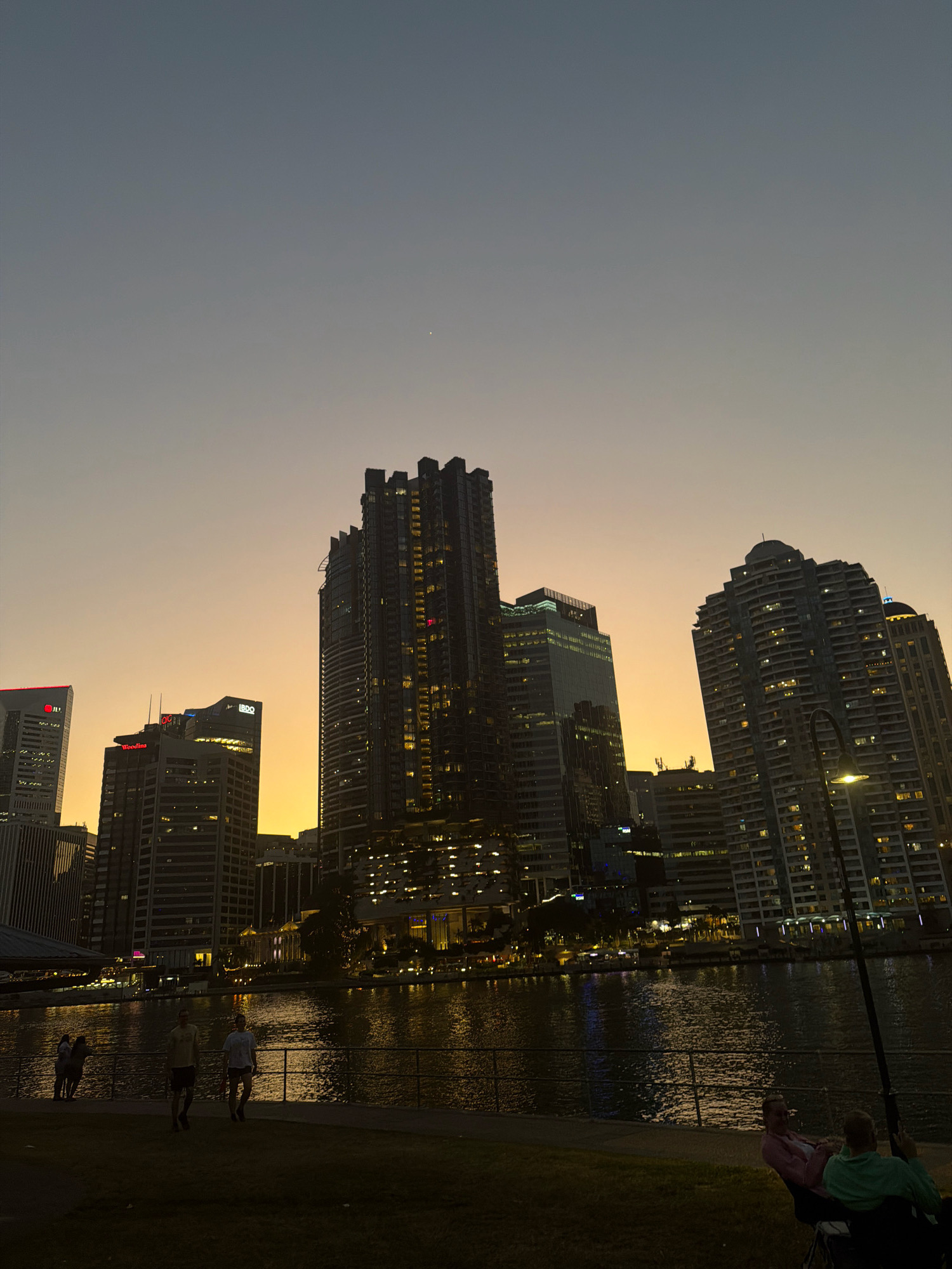 The image depicts a twilight scene of the Brisbane skyline viewed from Kangaroo Point, Australia. Tall, modern buildings with illuminated windows dominate the background, reflecting the warm hues of the setting sun in the sky, which transitions from a pale orange to a dusky blue. A wide river flows in the midground, its surface shimmering with the reflections of the city lights. Along the riverbank, a metal railing separates a paved walkway from a grassy area. Several individuals are present: on the walkway, a group of people are standing, and two individuals appear to be walking. In the foreground, on the grass, two people are seated in chairs, one wearing a pink shirt and the other a green shirt. A fishing rod is visible in the foreground. The overall atmosphere is tranquil, capturing the transition from day to night in an urban waterside setting.