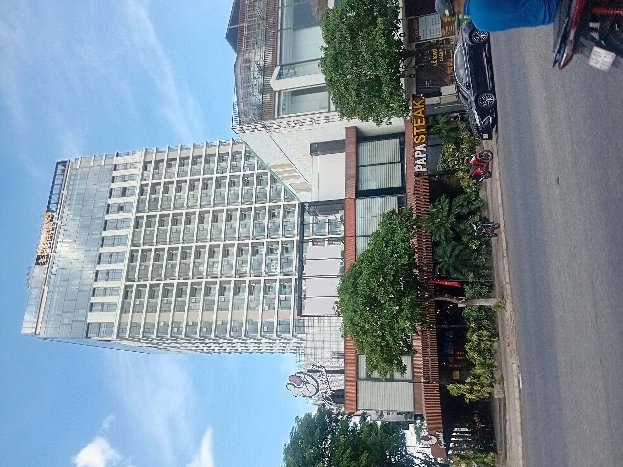 A tall, modern building with a glass facade and white vertical accents dominates the background under a blue sky with scattered clouds. The building has a prominent "LPBank" sign at its apex. In the foreground, a commercial building with a brown wooden slatted facade and large glass windows is visible, with "Papa Steak" signage on its exterior. Trees and greenery line the front of this lower building. A black car is parked on the street in front of Papa Steak, and several motorcycles are visible on the sidewalk and street. A person in a blue shirt is partially visible on the right side of the frame, facing away from the camera. The scene appears to be a street view in the city of Da Nang, Vietnam, likely in a commercial or business district.