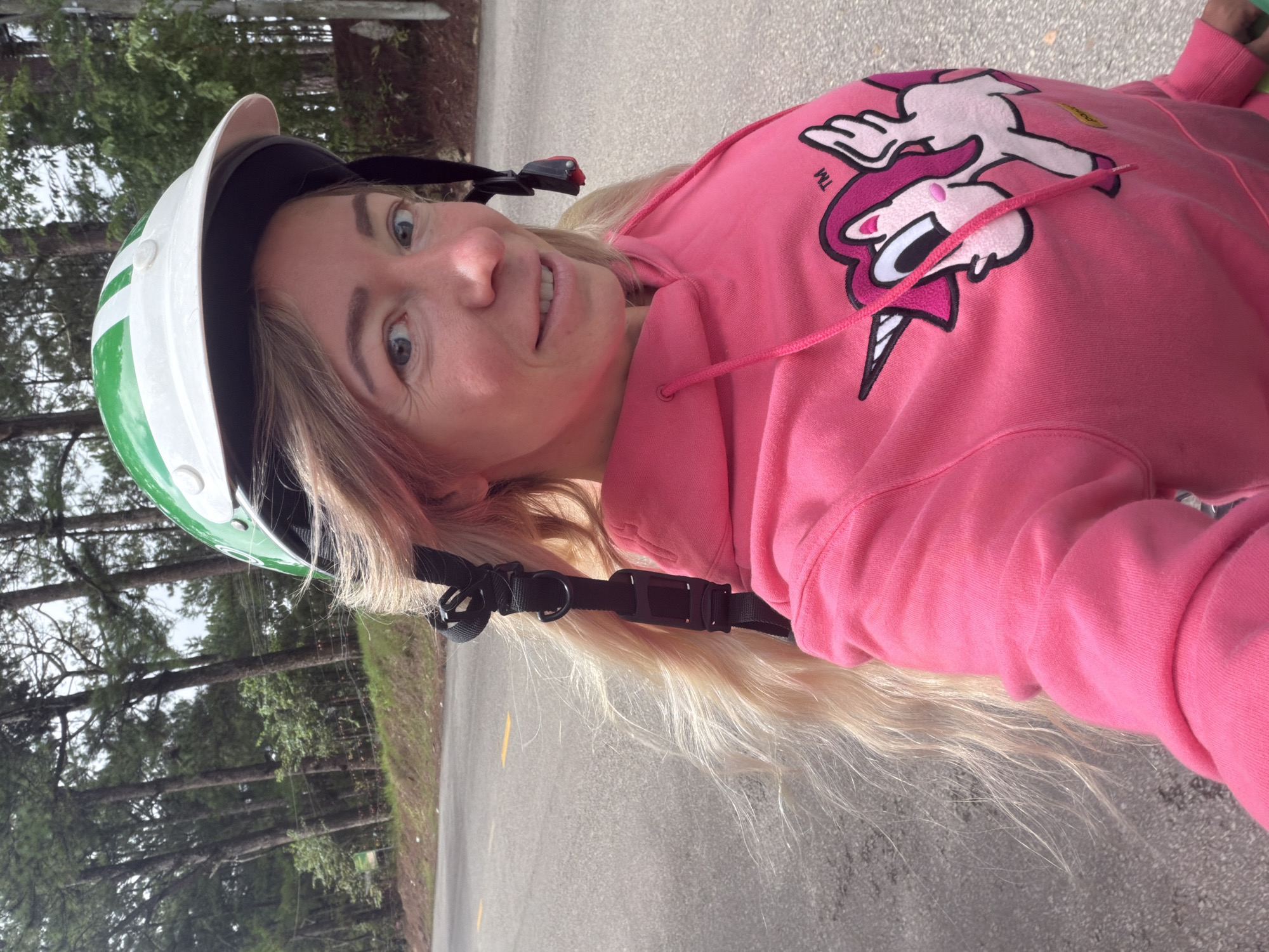 A blonde woman wearing a pink hoodie with a unicorn graphic and a green and white helmet is standing on a road, likely in Xuân Thành, Vietnam, given the context of the location. She is looking at the camera and smiling. The road is paved and has yellow lane markings. Pine trees line the side of the road, suggesting a rural or natural setting. A black strap from the helmet hangs down her hair, which is long and wavy. The woman is in the foreground, and the background consists of the road and trees.
