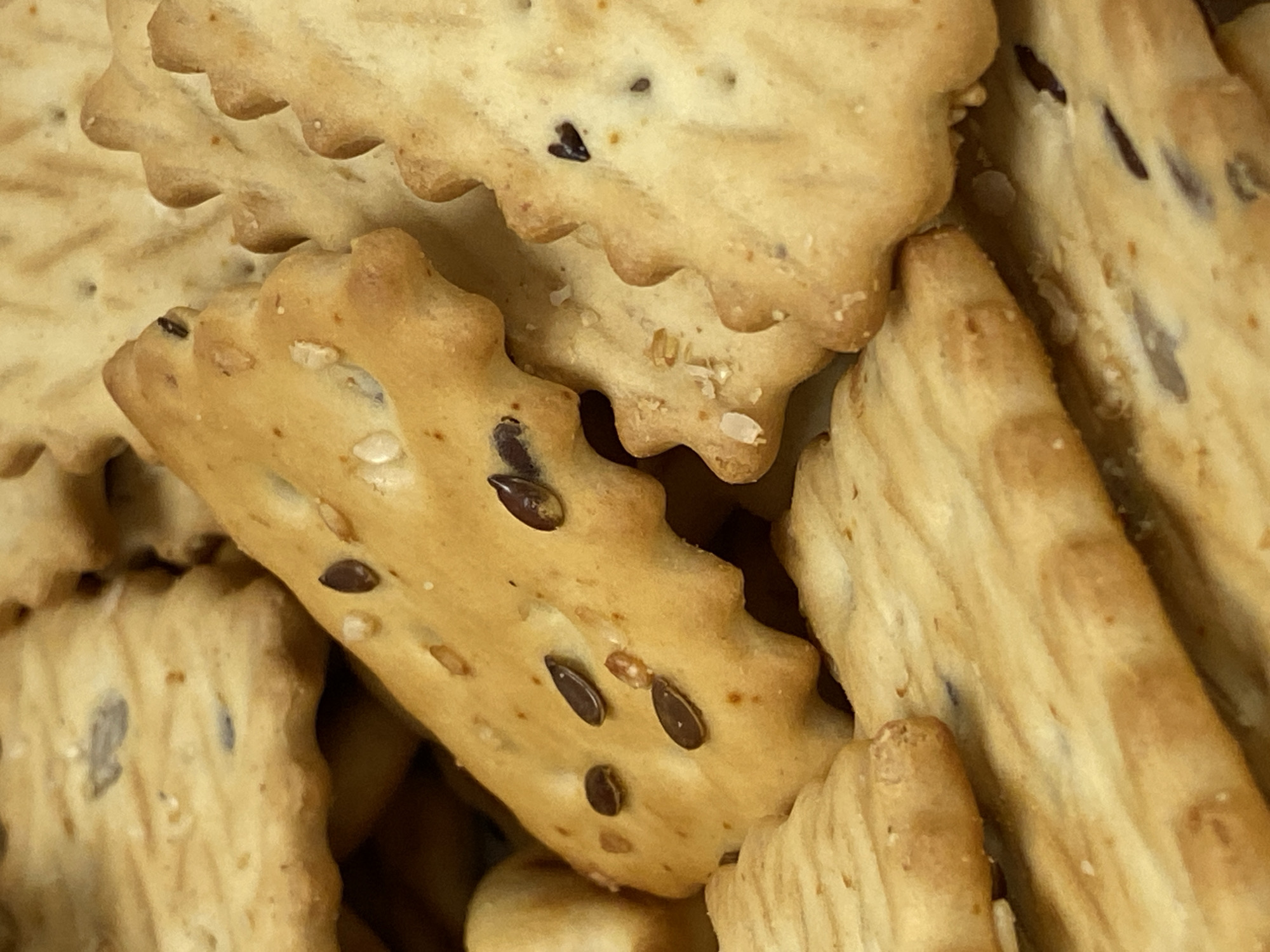 A close-up image shows a cluster of flaxseed crackers. The crackers have a light golden-brown color with visible flax seeds embedded in their surface. Some of the crackers have decorative, slightly scalloped edges. The environment appears to be indoors. The location is not discernible and cannot be determined to be Votkinsk, Russia, based on the visual information provided.