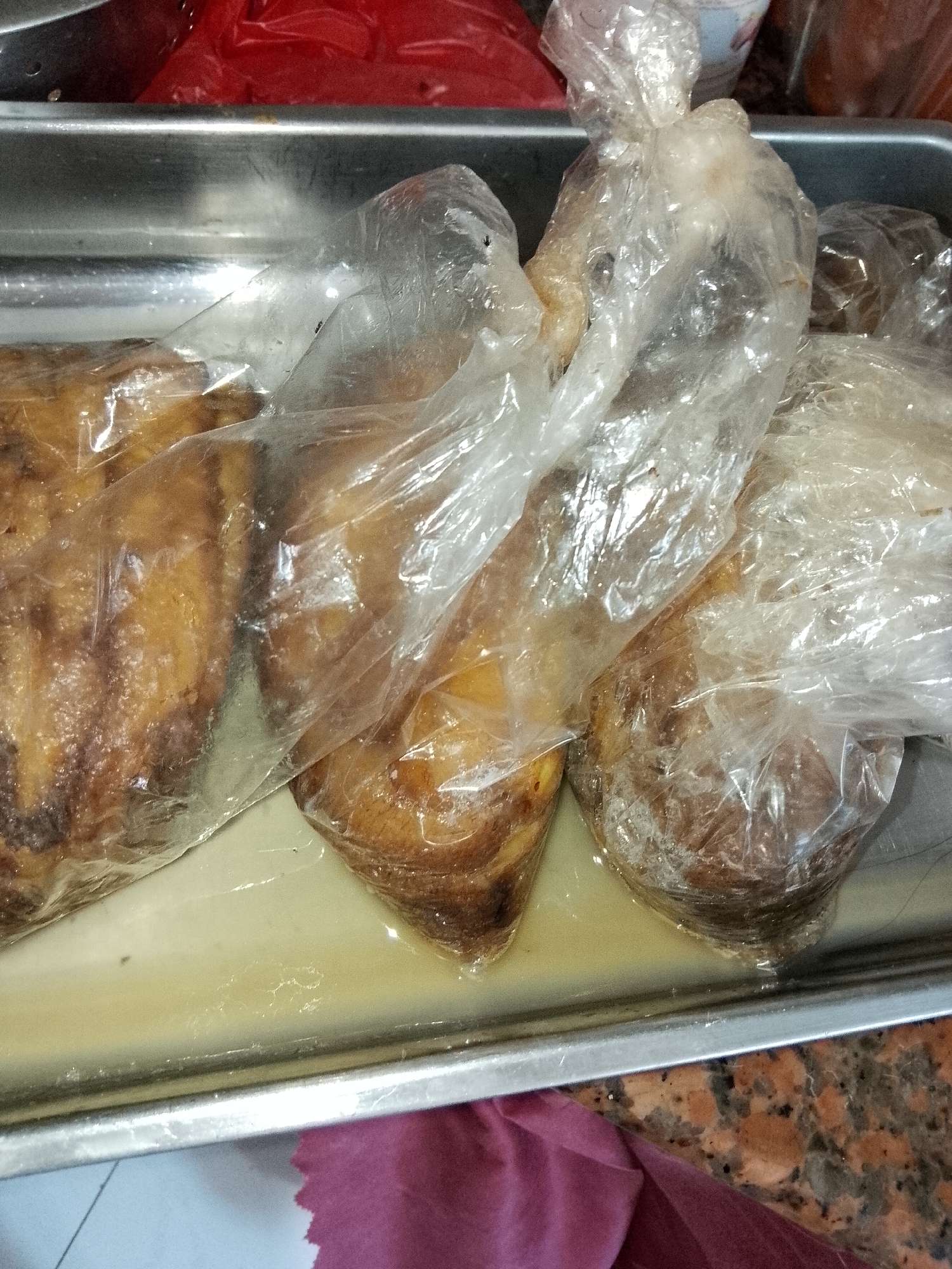 The image shows several food items, likely baked goods or fried snacks, individually wrapped in clear plastic bags and placed in a shallow metal tray. The food has a golden-brown, somewhat irregular texture, suggesting items such as bread, cakes, or possibly fried plantains or fritters. A pale yellow, translucent liquid, possibly oil or syrup, has collected at the bottom of the tray, with some of the wrapped items partially submerged in it.
The metal tray is utilitarian, reflecting light on its surface. In the background, out of focus, are objects in red plastic bags or containers, along with a glimpse of another container with a white and blue pattern. The scene is set indoors, suggesting a kitchen, bakery, or food preparation area. A piece of pink or maroon fabric is visible in the lower right corner, resting on a multi-toned, possibly granite, countertop.
No people are visible in the image. The activity depicted appears to be the packaging or temporary storage of prepared food items. There is no visible text. The overall atmosphere is functional and practical, focused on food handling.