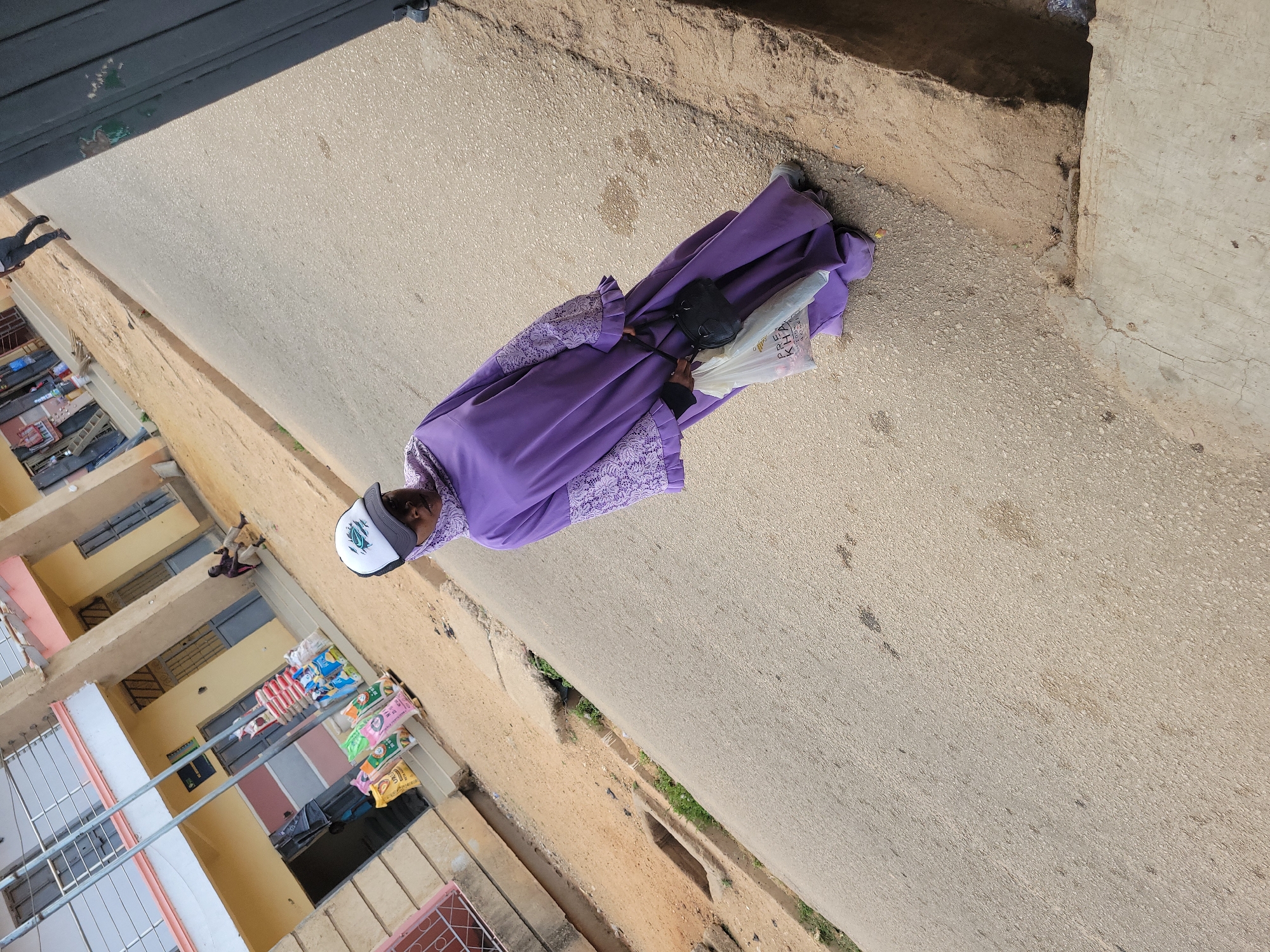 A woman wearing a long purple dress and a white baseball cap is walking on a paved street in Jos, Nigeria. She is carrying a black purse and a white plastic bag. The street is lined with buildings. Laundry is hanging on the line outside one of the buildings.