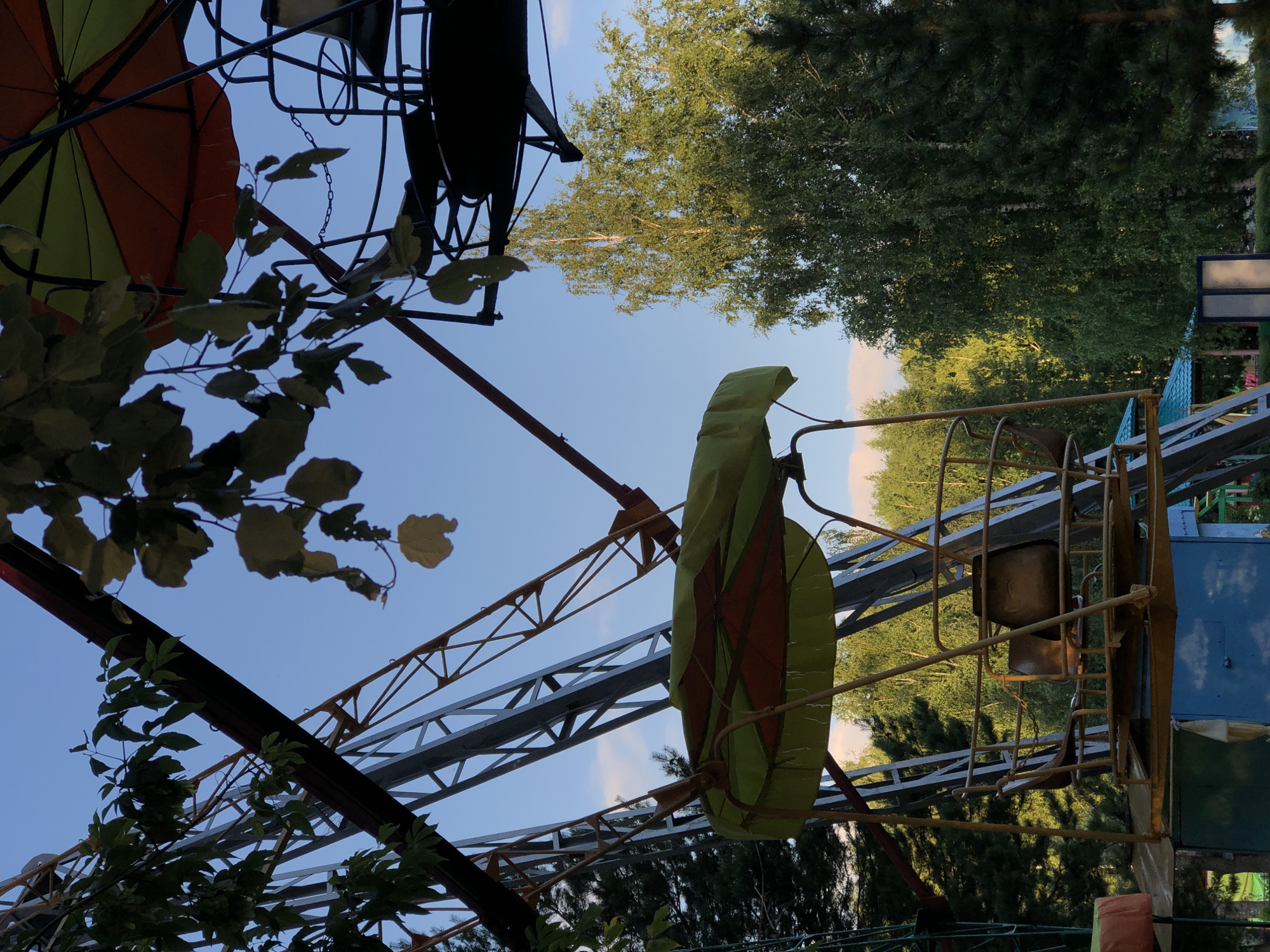 This media file captures a close-up, low-angle view of an amusement park ride, possibly a Ferris wheel or a similar structure, against a clear blue sky. The perspective is tilted, with parts of the ride and surrounding foliage dominating the foreground.
Visible objects include:
- A brightly colored umbrella, divided into orange and yellow sections, partially obscuring the sky.
- Several metal support beams and frameworks of the ride, painted in dark colors, rust, and yellow.
- Green leaves and branches from nearby trees or vines are intertwined with the ride's structure.
- A weathered, yellow-green fabric covering, possibly a part of a ride car or canopy, is prominent.
- The hint of seats or gondolas within a cage-like structure suggests it's a ride designed to carry passengers.
- A blue panel with a white rectangle and a green panel are also visible on the right side.
The scene is outdoors, with abundant greenery from trees in the background, suggesting a park or fairground setting. The clear blue sky indicates fair weather and likely daytime, possibly late afternoon given the lighting. No people are visible, and no specific activity or event is taking place; it appears to be a still shot of the ride's infrastructure. The overall mood is somewhat rustic, with visible signs of wear on the metal structures. No text is discernible in the image. The location context is Chaikovsky, Russia.