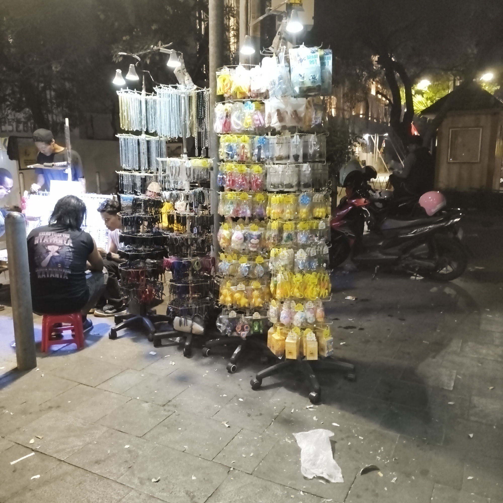 This image depicts a street stall in West Jakarta, Indonesia, at night. The stall displays various accessories, including necklaces, keychains, and small packaged items arranged on racks. A vendor is visible behind the stall, interacting with customers. Two men are seated on stools in front of the stall, one of whom is wearing a black t-shirt with text on the back. A motorcycle with a pink helmet is parked to the right of the stall. The ground is paved with stone tiles and is littered with debris, including paper and plastic. The scene is illuminated by artificial lights from the stall and surrounding areas, casting shadows on the ground.