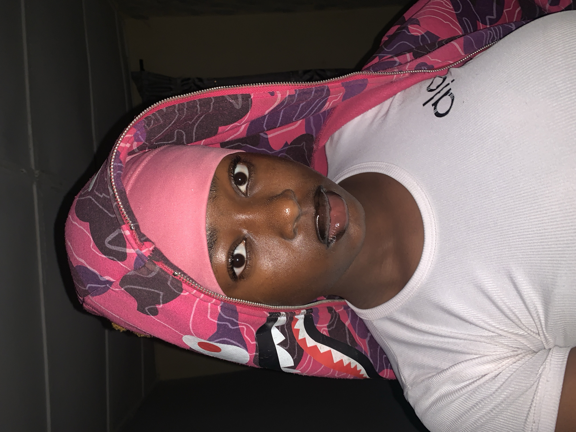 A close-up, indoor shot shows a dark-skinned person wearing a pink headscarf, a white tank top with the logo "Alo", and a pink and purple camo jacket. The jacket's hood is up, framing the person's face, and features a shark-like design near the hood's opening. The person's mouth is open. The location is not discernible as Rafin Par, Nigeria, from the image. The background is a dark, indistinct wall.