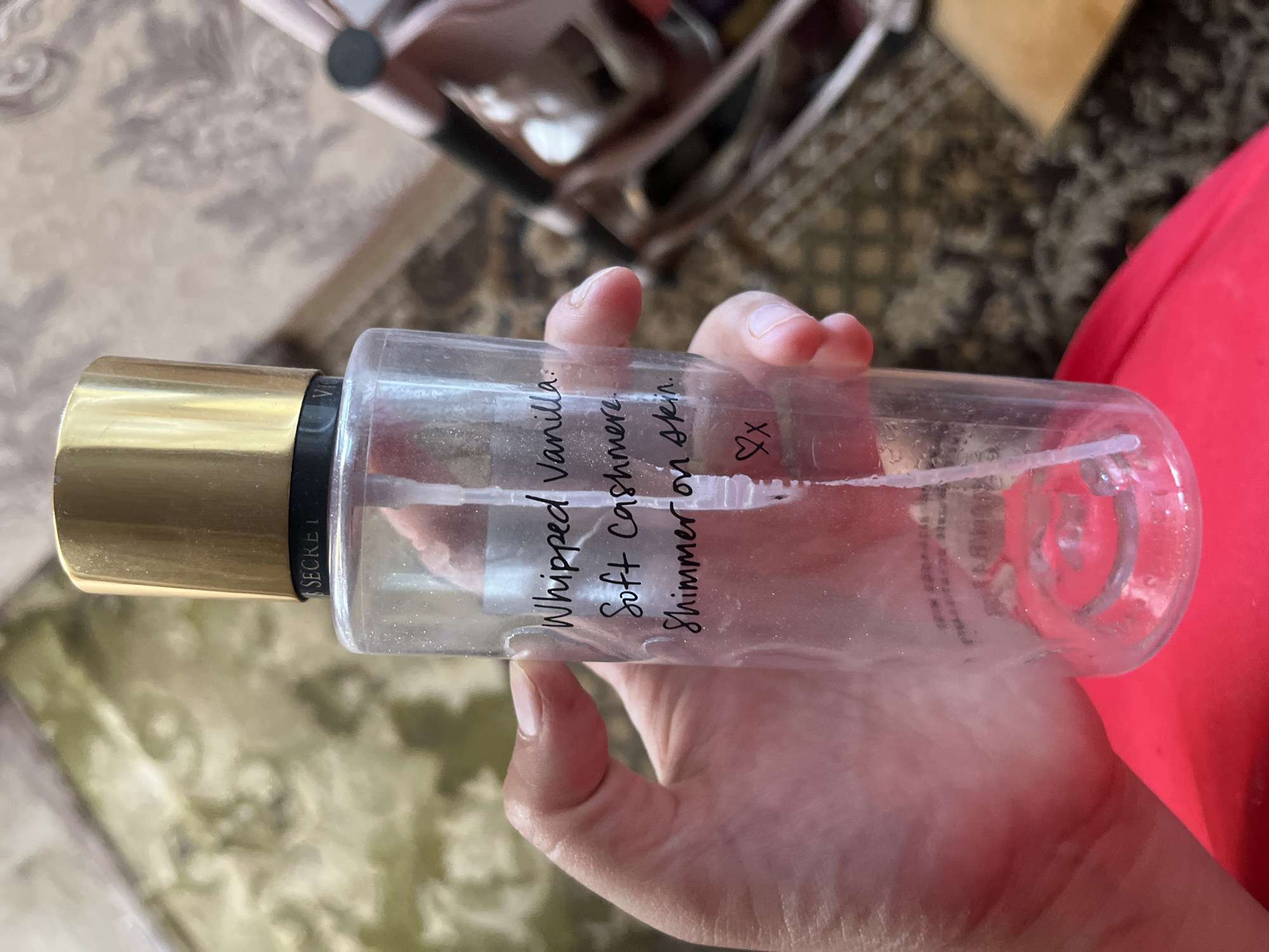 A hand holds a nearly empty, clear plastic bottle with a gold cap. The bottle has black handwritten text that reads "Whipped Vanilla: Soft Cashmere. Shimmer on skin. ♡x". The background is blurred and appears to be an indoor setting with a patterned rug and furniture. The lighting suggests it is daytime.