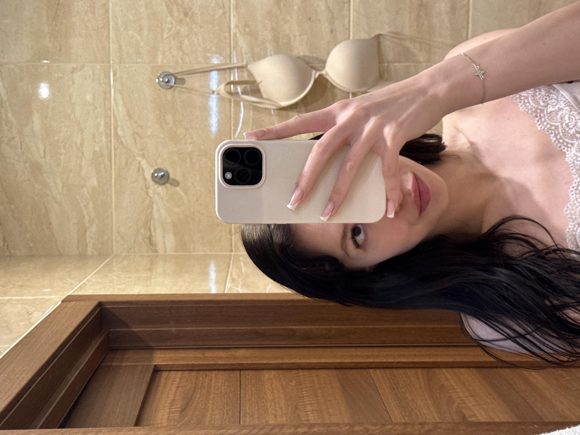 A woman is taking a selfie in a bathroom. She is holding a smartphone in her right hand and is partially covering her face with it. She has long, dark hair and is wearing a light-colored lace-trimmed top. Her left arm is visible, and she is wearing a bracelet with a star charm. In the background, there are beige tiled walls and a wooden doorframe. A beige bra is hanging on hooks on the wall behind her. The scene appears to be a private residence.