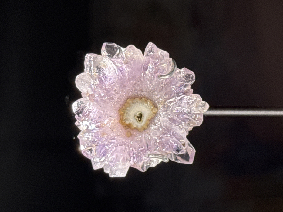 A crystal flower with an amethyst hue dominates the image against a black background. The crystal appears to be constructed from many pointed shards, giving it a textured, petal-like form. The flower's center exhibits a light tan color, contrasted by a small, circular white core with a dark spot in the center. The presentation of the flower implies it is on display, possibly for viewing or examination.