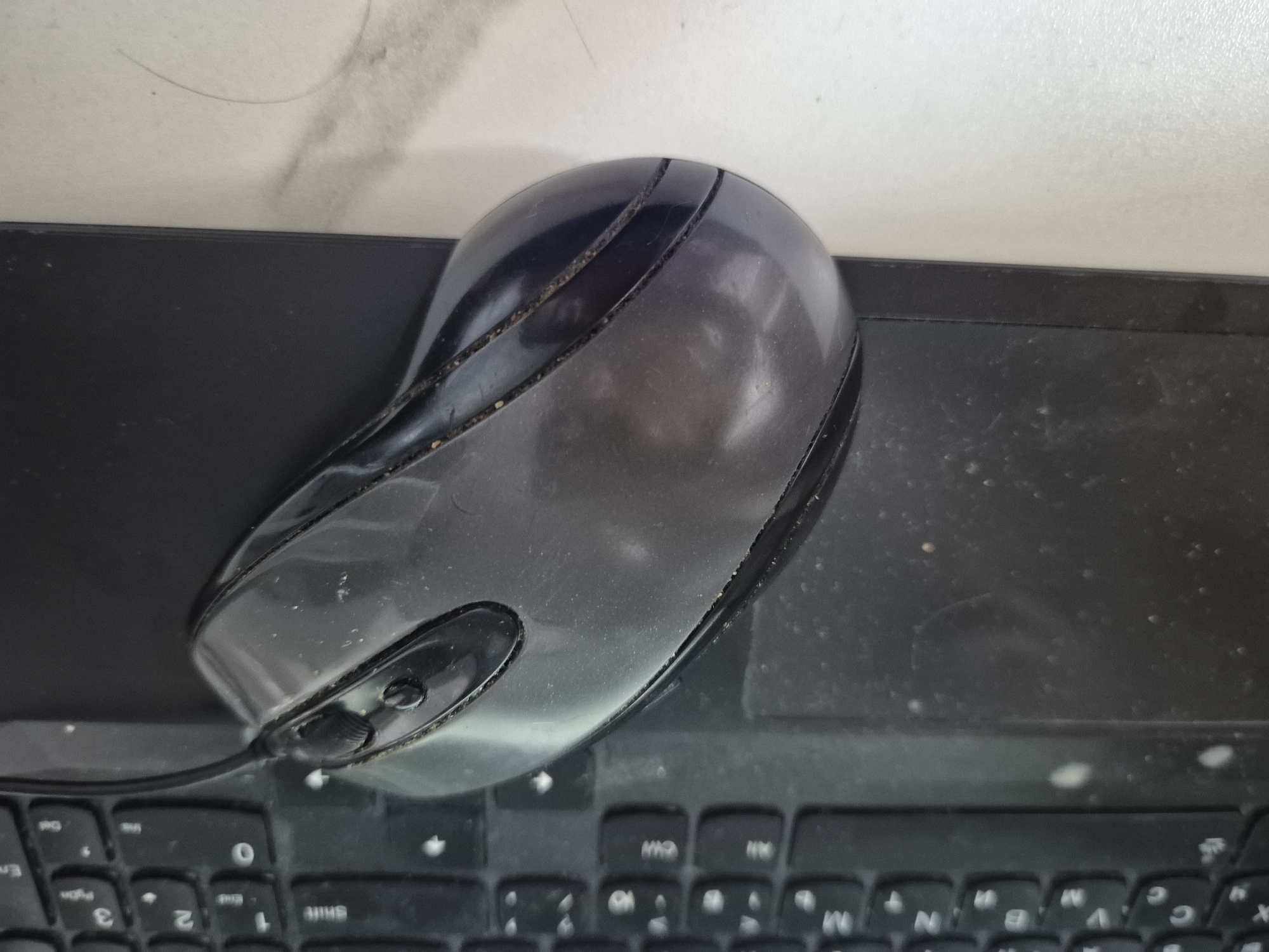 The image shows a close-up, slightly angled view of a black computer mouse positioned on top of a black keyboard. The mouse is translucent and appears to be made of plastic, with visible dust and grime on its surface and along the seams. It has two buttons on top and a scroll wheel in between them. The keyboard is also black and appears to be a laptop keyboard, with some keys visible, including numbers and letters like 'Shift', 'End', '3', '2', '1', and '0'. The background to the right of the keyboard is a light-colored surface, possibly a desk or table, which also shows some smudges and dust. The lighting suggests an indoor setting. There are no people or animals in the image, and no specific actions or interactions are depicted. The image does not provide any information about the location in the city of Astana, Kazakhstan.